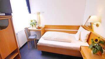 A hotel room with a single bed, a desk with a chair, a lamp, a radiator, and a television.
