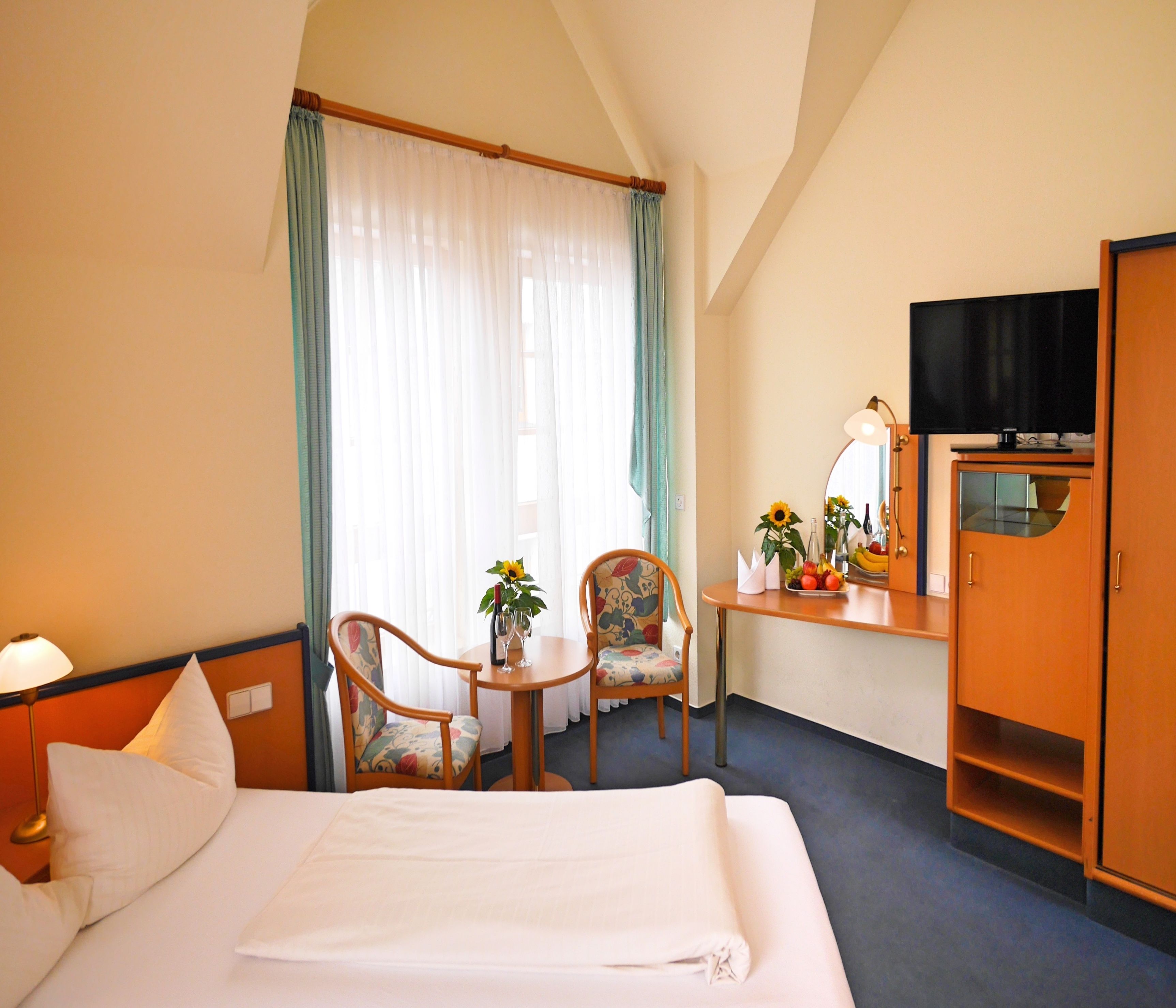 A hotel room with a bed, two chairs, a small table with a vase of flowers, a desk with a mirror, and a television on a cabinet.