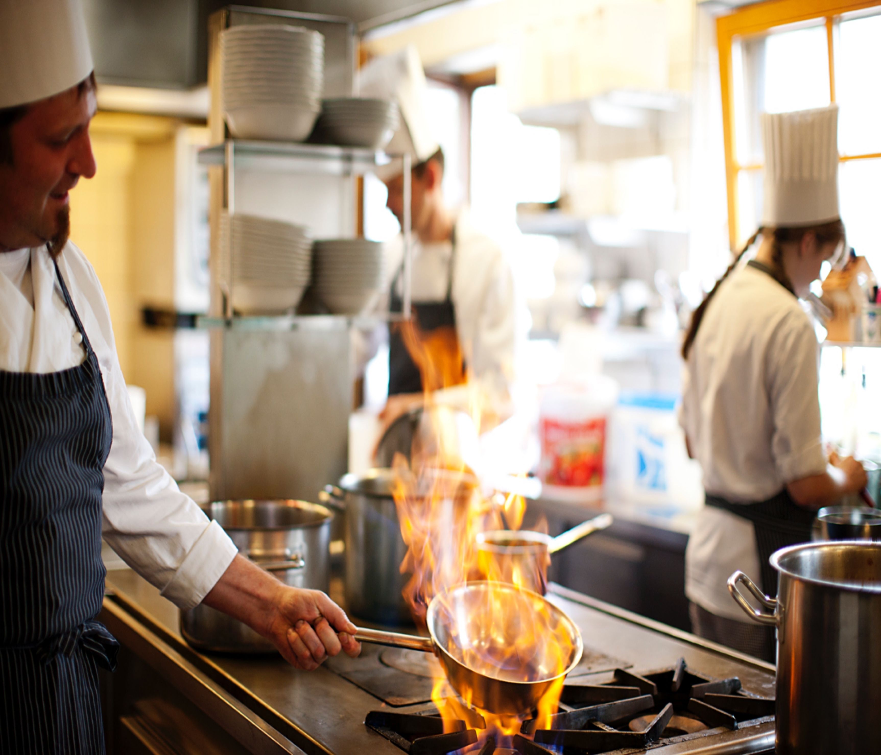 A kitchen with chefs cooking, stainless steel pots, and a stove with flames.