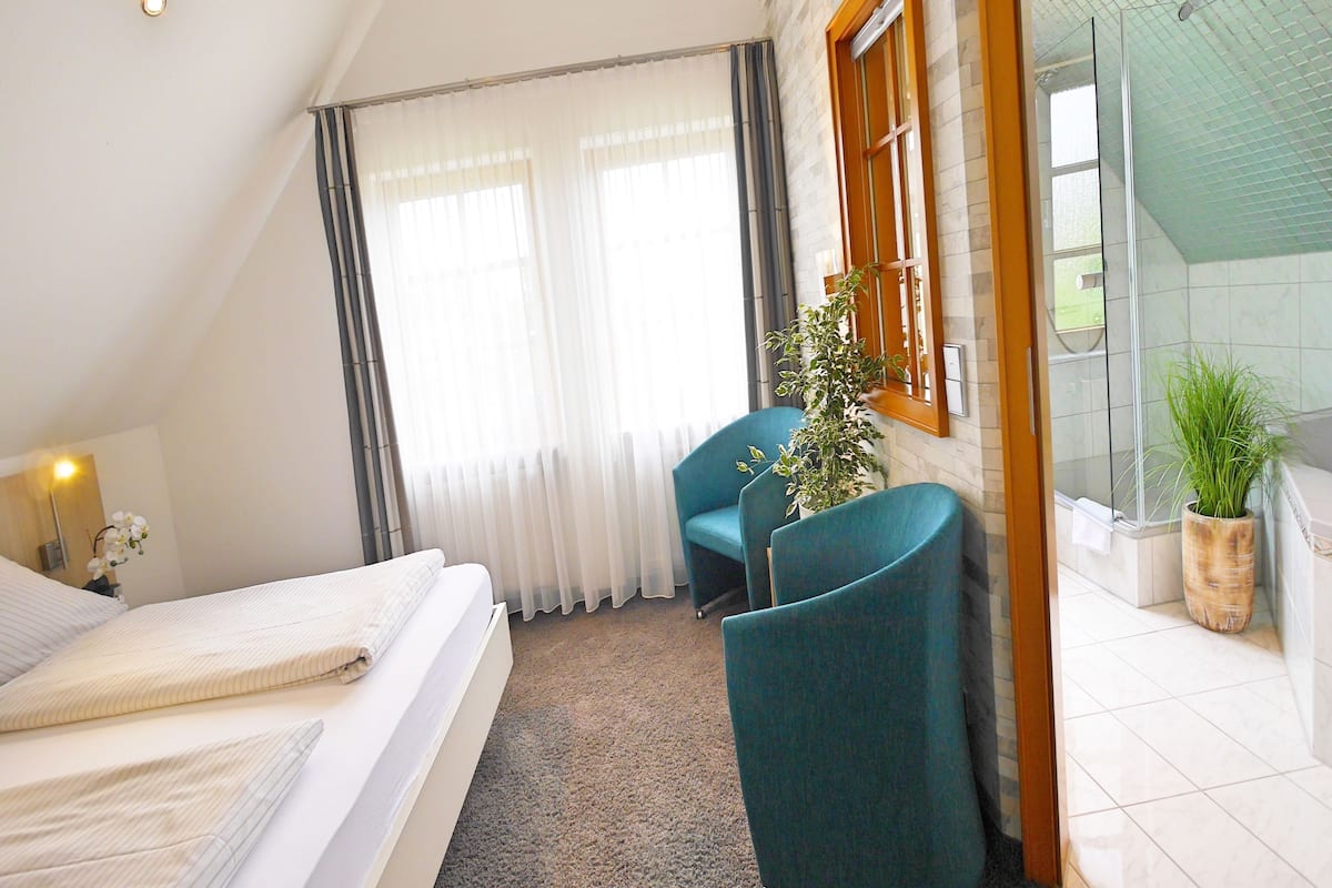 Een slaapkamer met een bed, een bureau, een stoel, een kamerplant en een badkamer met een glazen deur.