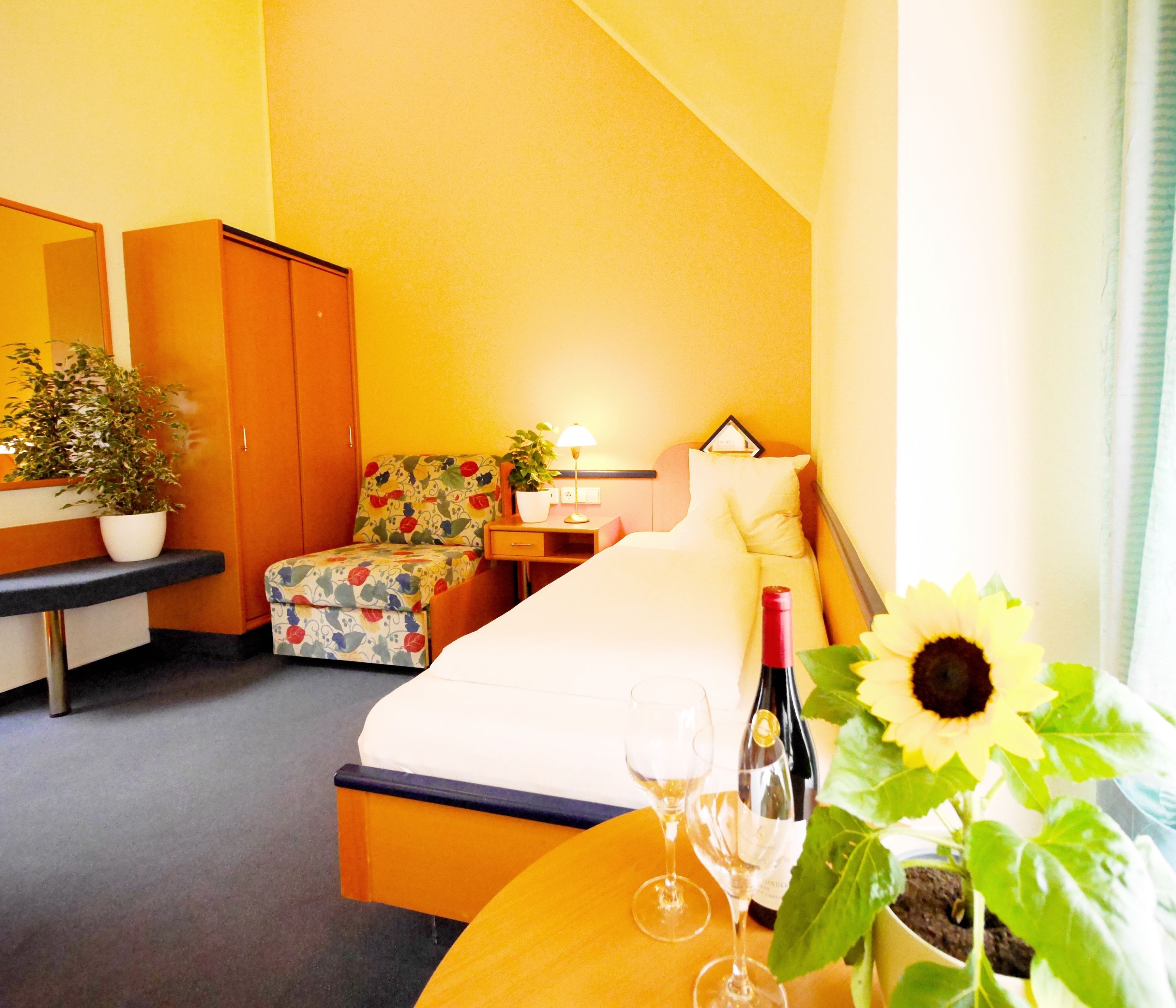 A hotel room with a bed, a desk, a chair, a nightstand, a lamp, a potted plant, a wine bottle, and a glass of wine.