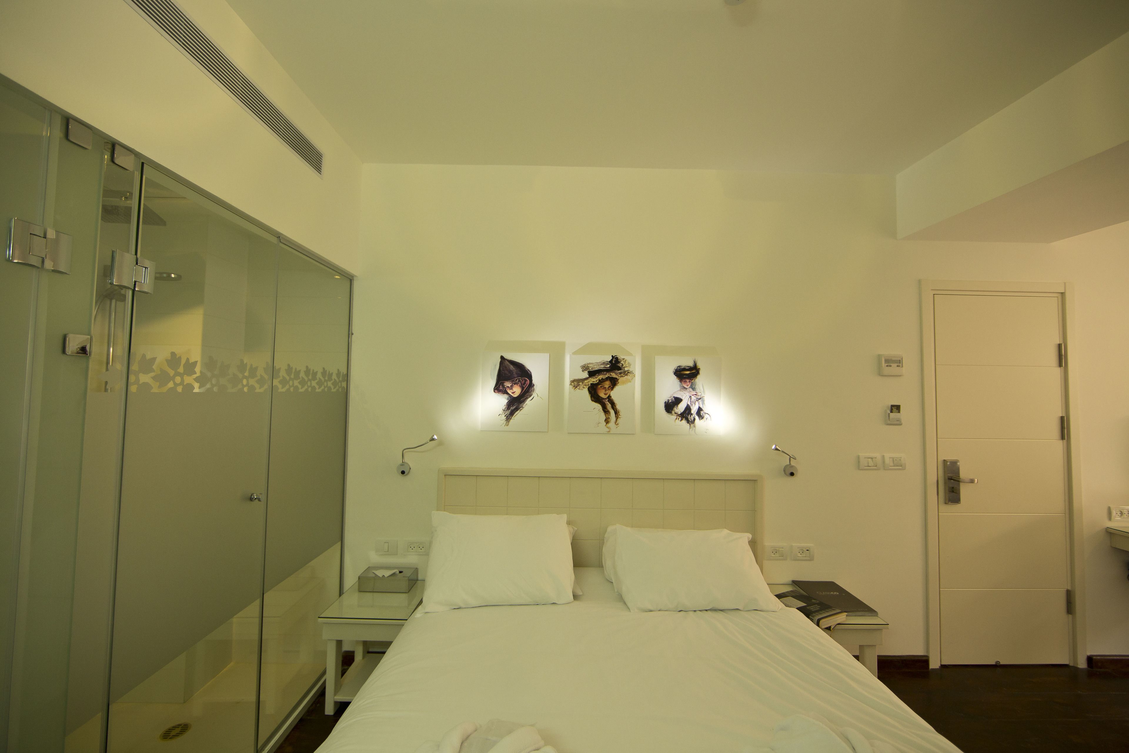 A hotel room with a bed, bedside table, and a wardrobe. There are wall-mounted lights and a door with a handle.