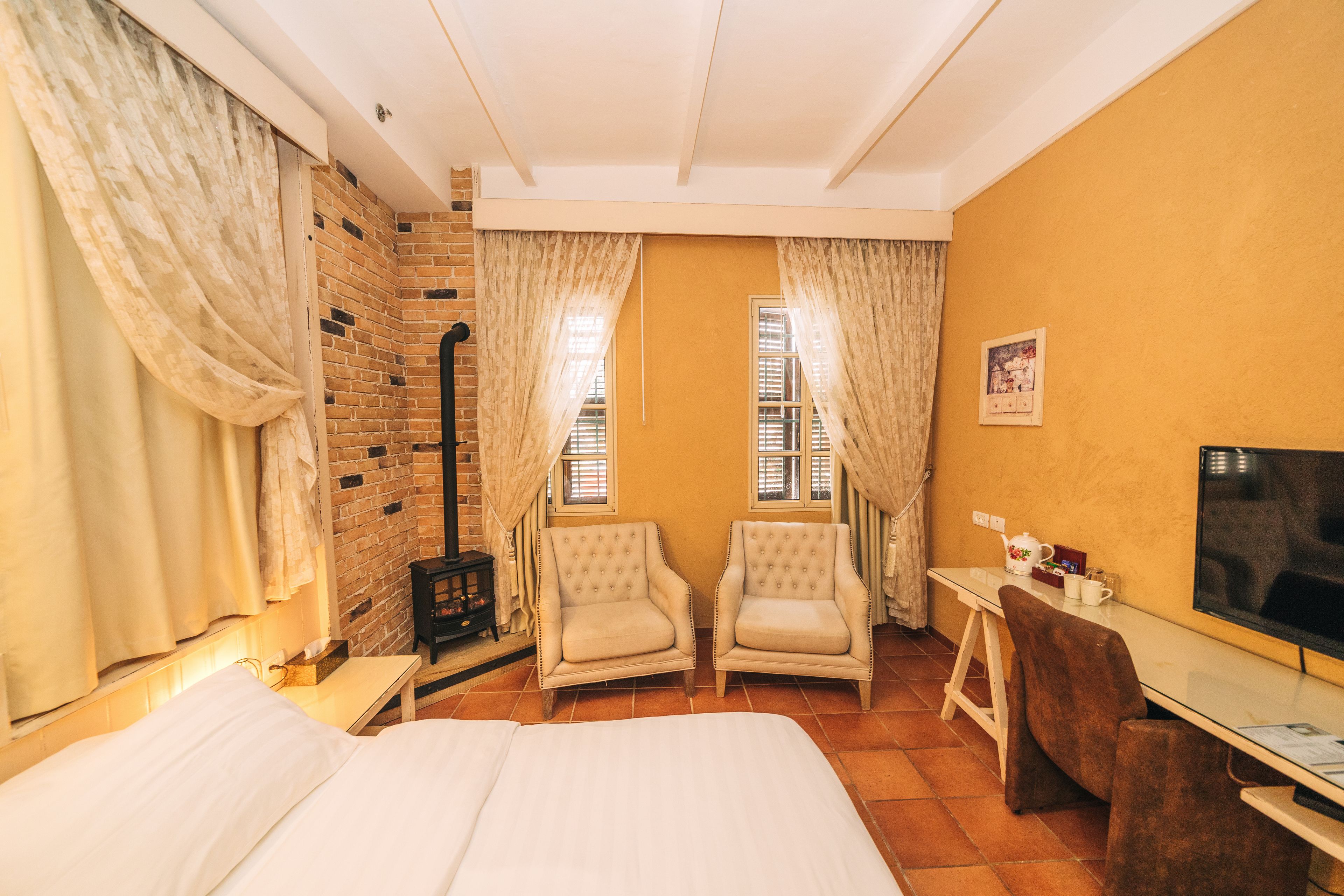 A hotel room with a bed, two armchairs, a small table, a TV, and a brick wall.