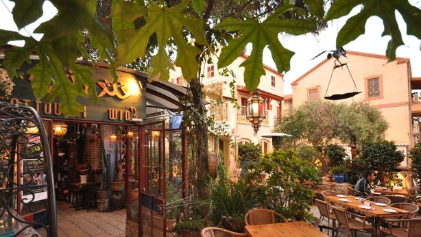 Um café ao ar livre com mesas e cadeiras de madeira, cercado por plantas em vasos e um edifício com uma placa.