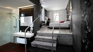Un baño moderno con ducha de vidrio, lavamanos y una escalera que conduce a un dormitorio con cama y mesita de noche.
