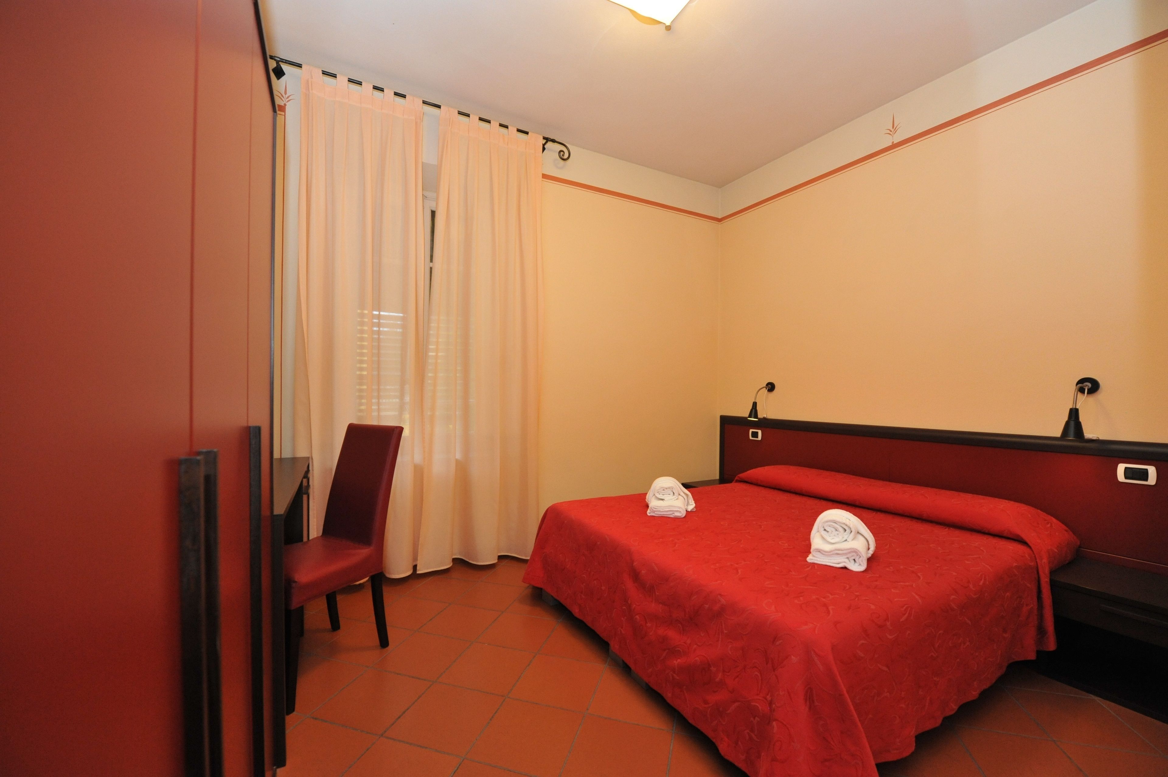 A hotel room with a red bed, a chair, a desk, and a window with curtains.