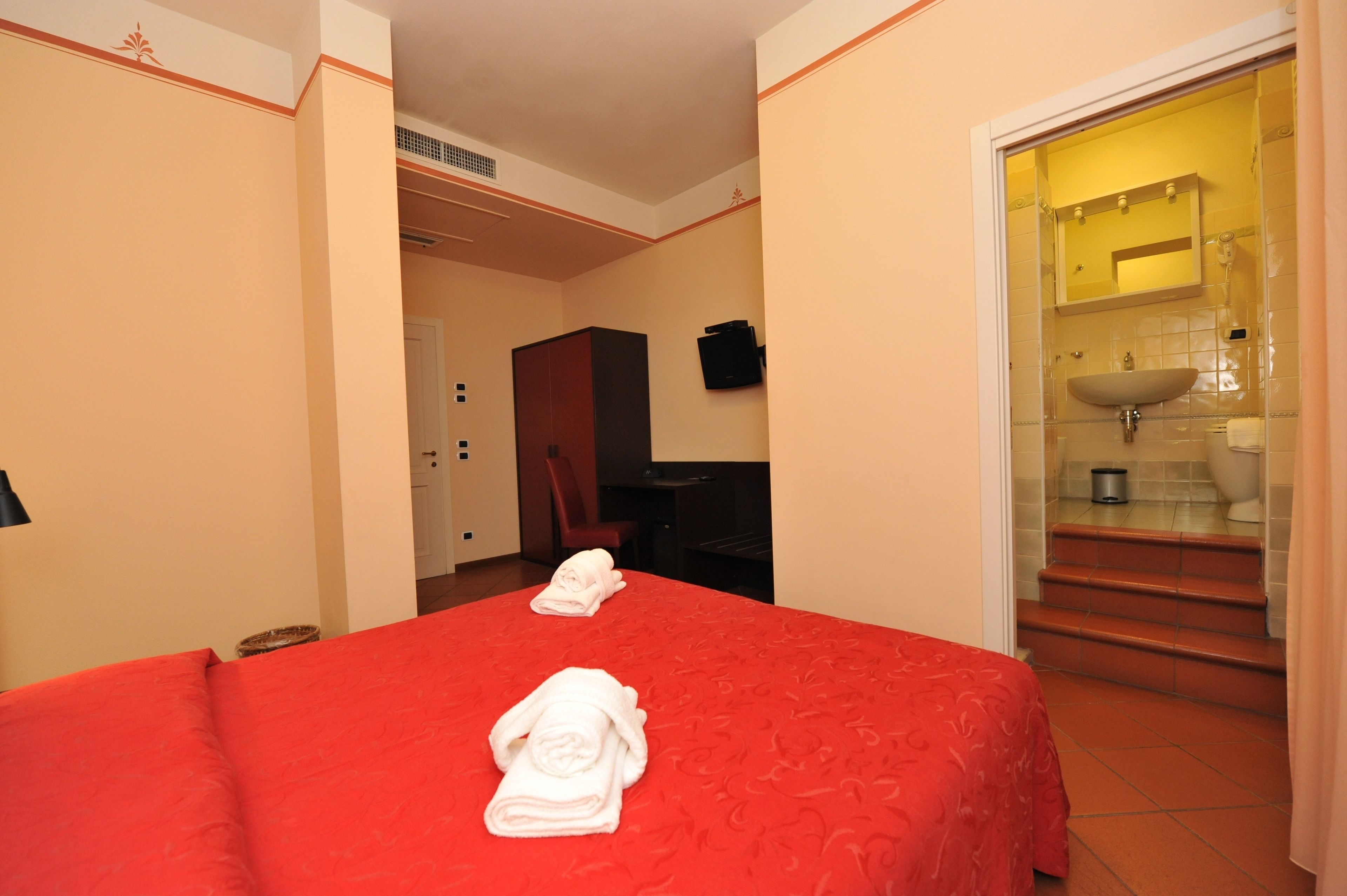 A hotel room with a red bedspread, a neatly folded towel, a desk with a chair, and a bathroom with a sink and mirror.