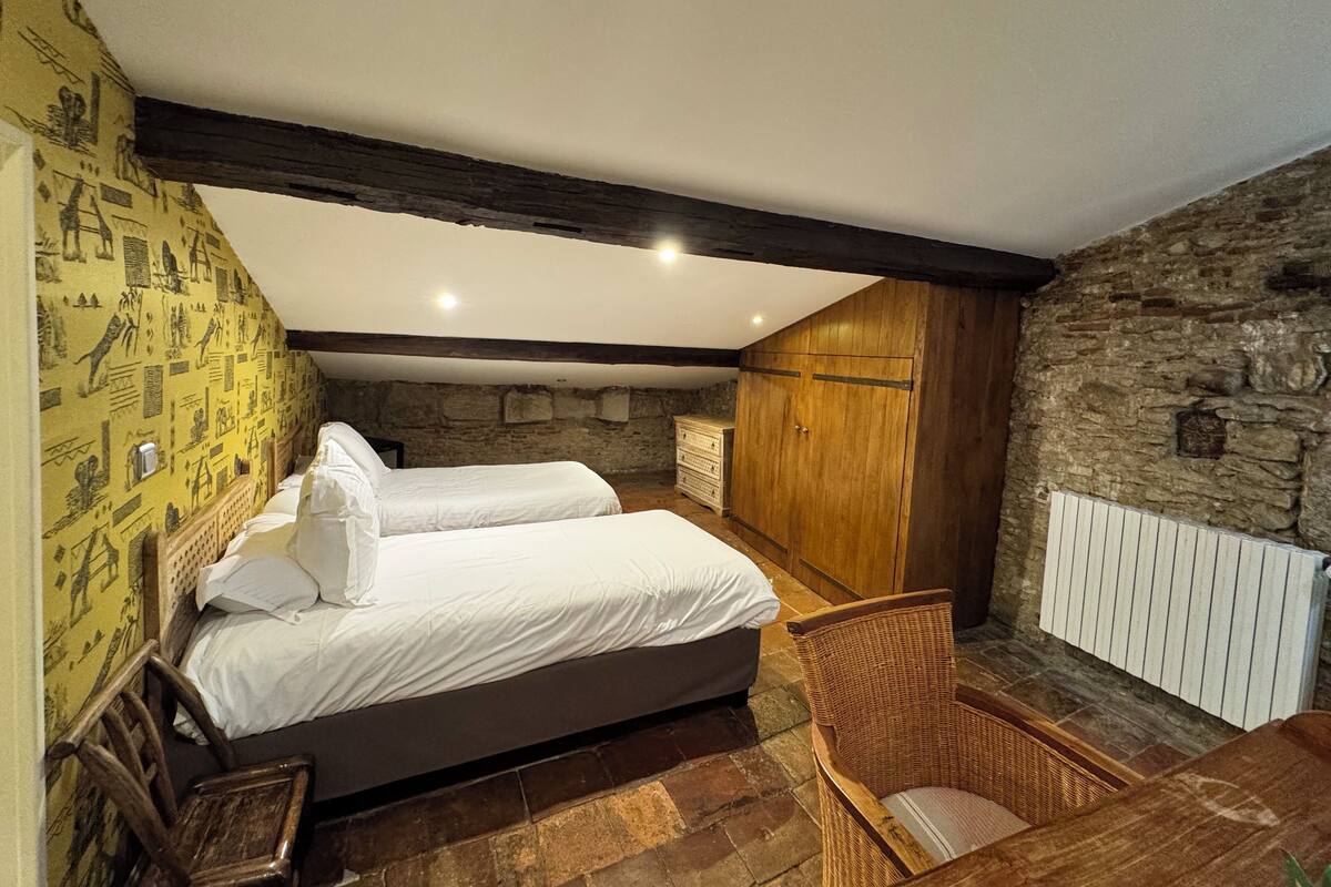 A bedroom with two beds, a stone wall, a wooden wardrobe, and a wicker chair.