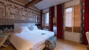 Une chambre avec un grand lit, un banc à motifs zébrés, un mur décoré de fresques représentant des animaux et des plantes, et une fenêtre avec des stores.