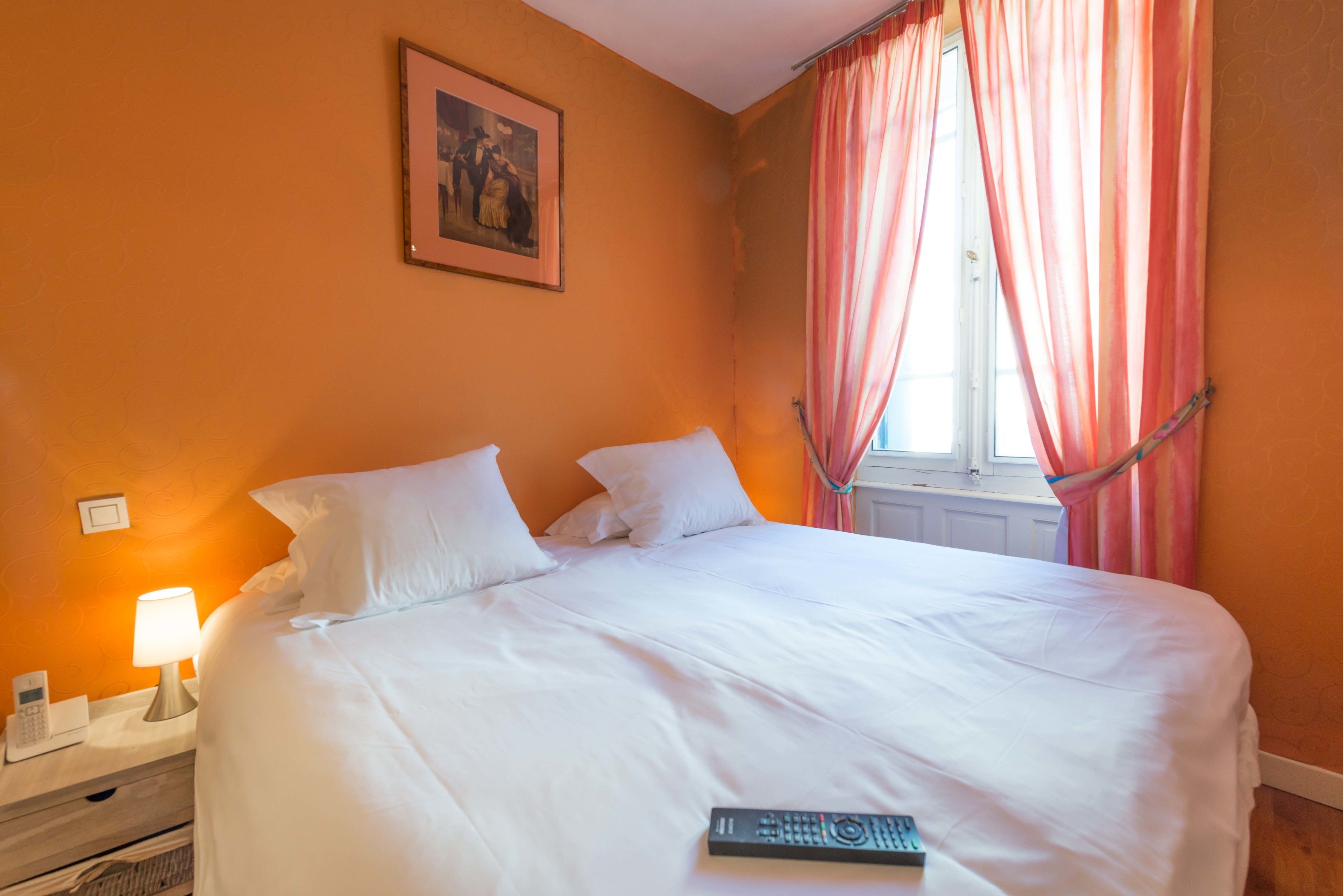 A neatly made bed with white linens, a bedside lamp, a framed picture on the wall, and a window with pink curtains.