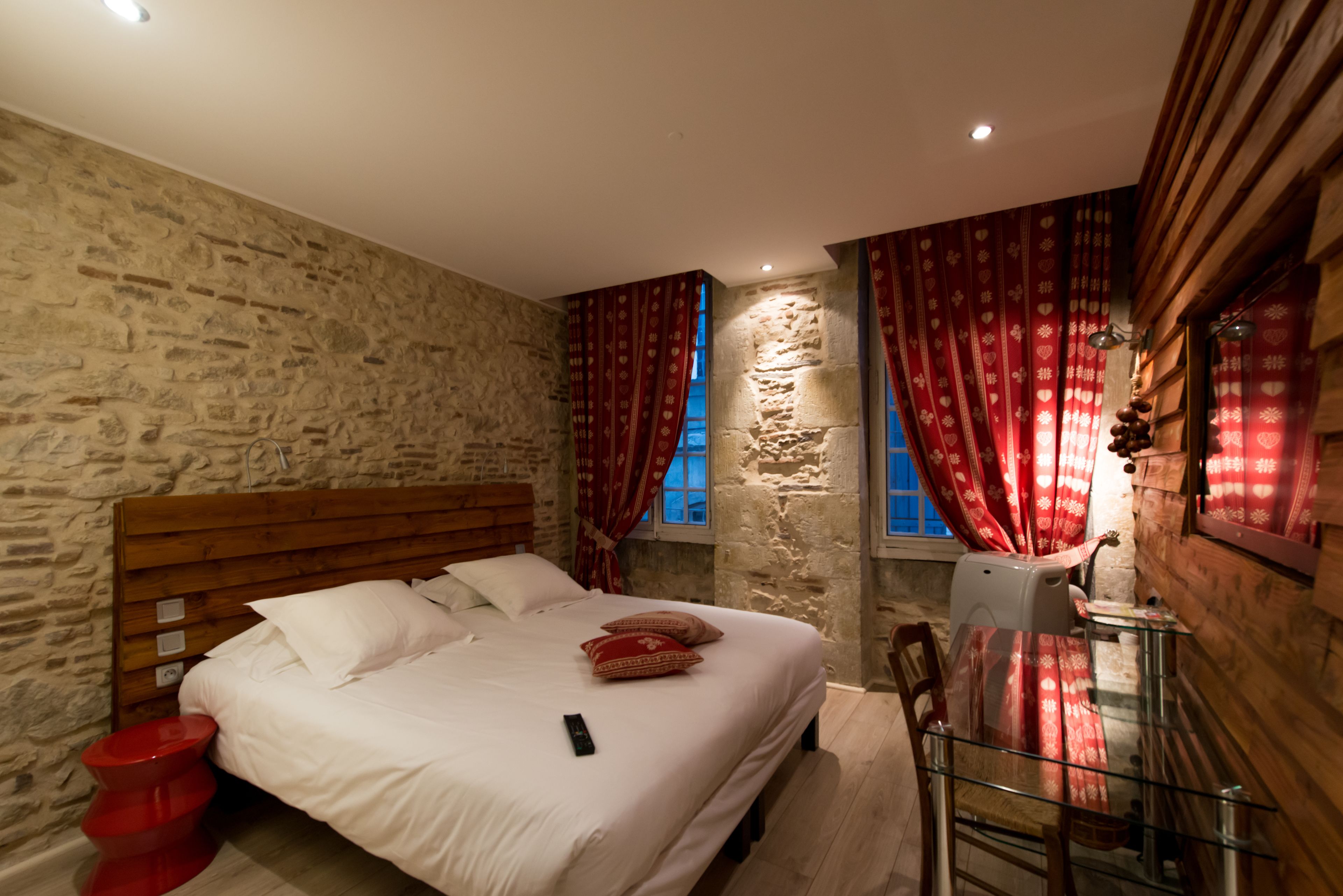 Un dormitorio con pared de piedra, cabecero de madera, mesa de vidrio y cortinas rojas.