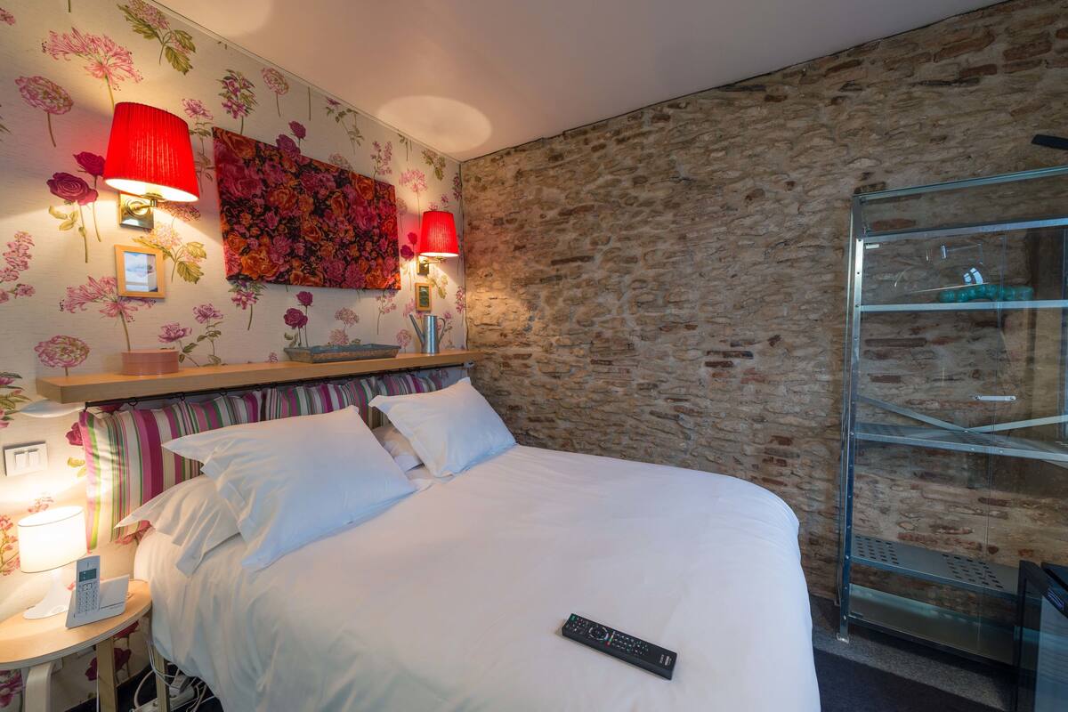 A hotel room with a stone wall, a bed with white linens, a nightstand with a lamp, and a glass shelving unit.