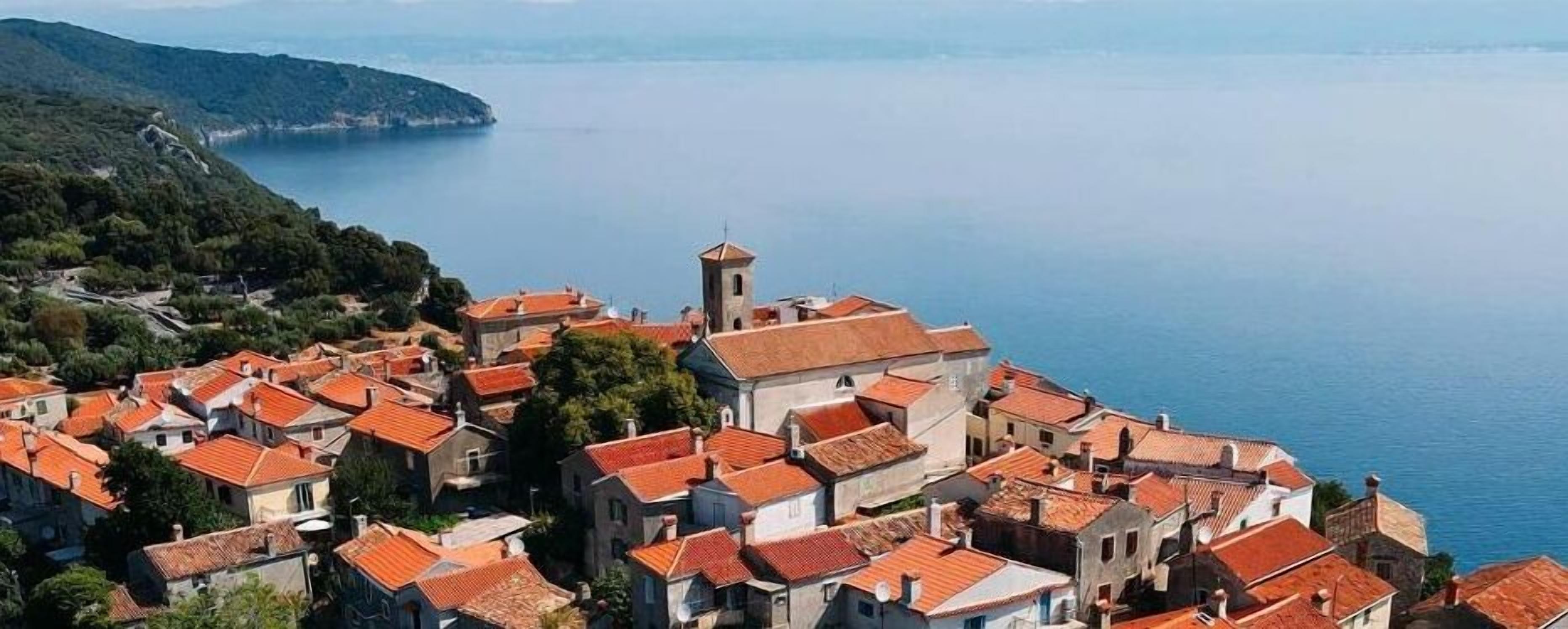 A coastal village with terracotta-roofed buildings, a prominent tower, and a view of the sea.