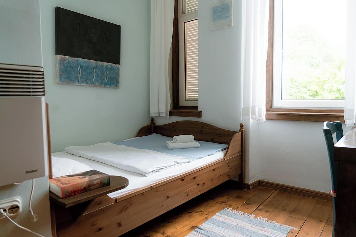 A single bed with a wooden frame, a bedside table with a book, a window with blinds, and a wall-mounted heater.