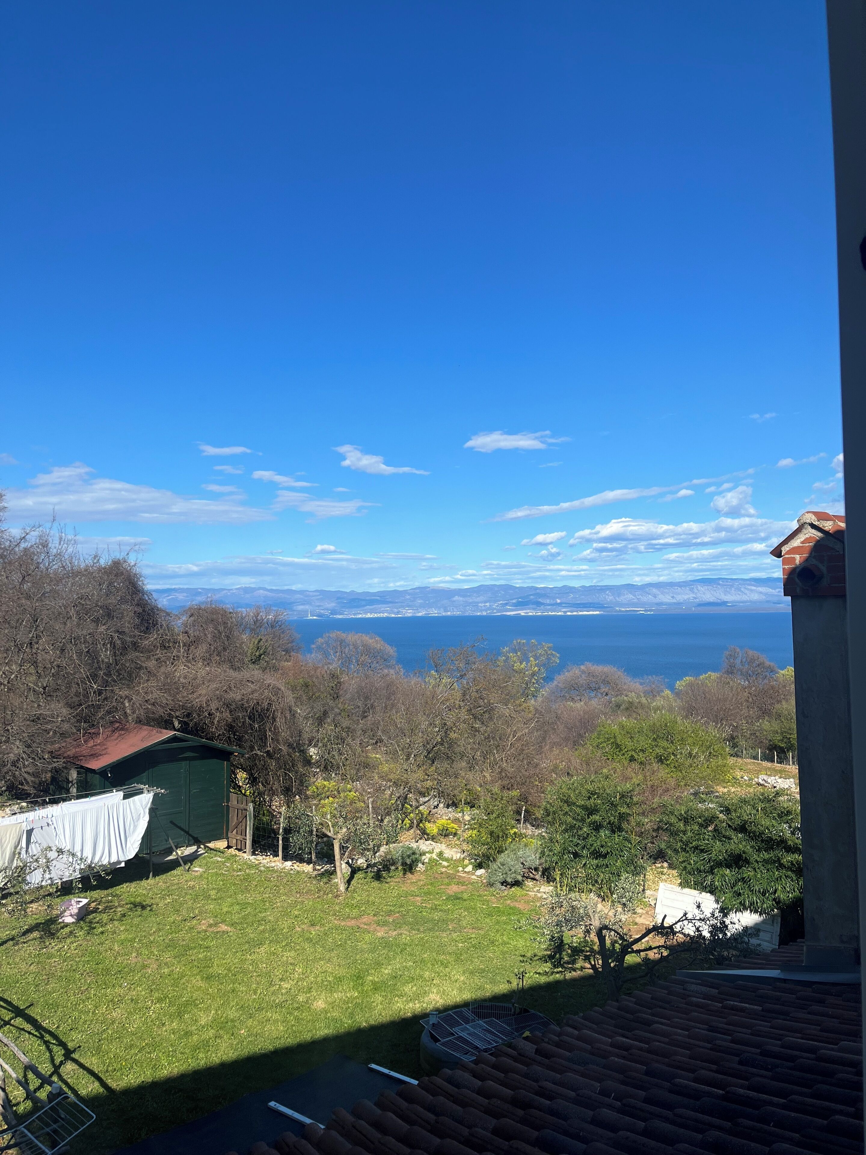 A view from a balcony overlooking a grassy yard with a small shed, a metal fence, and a body of water in the distance.