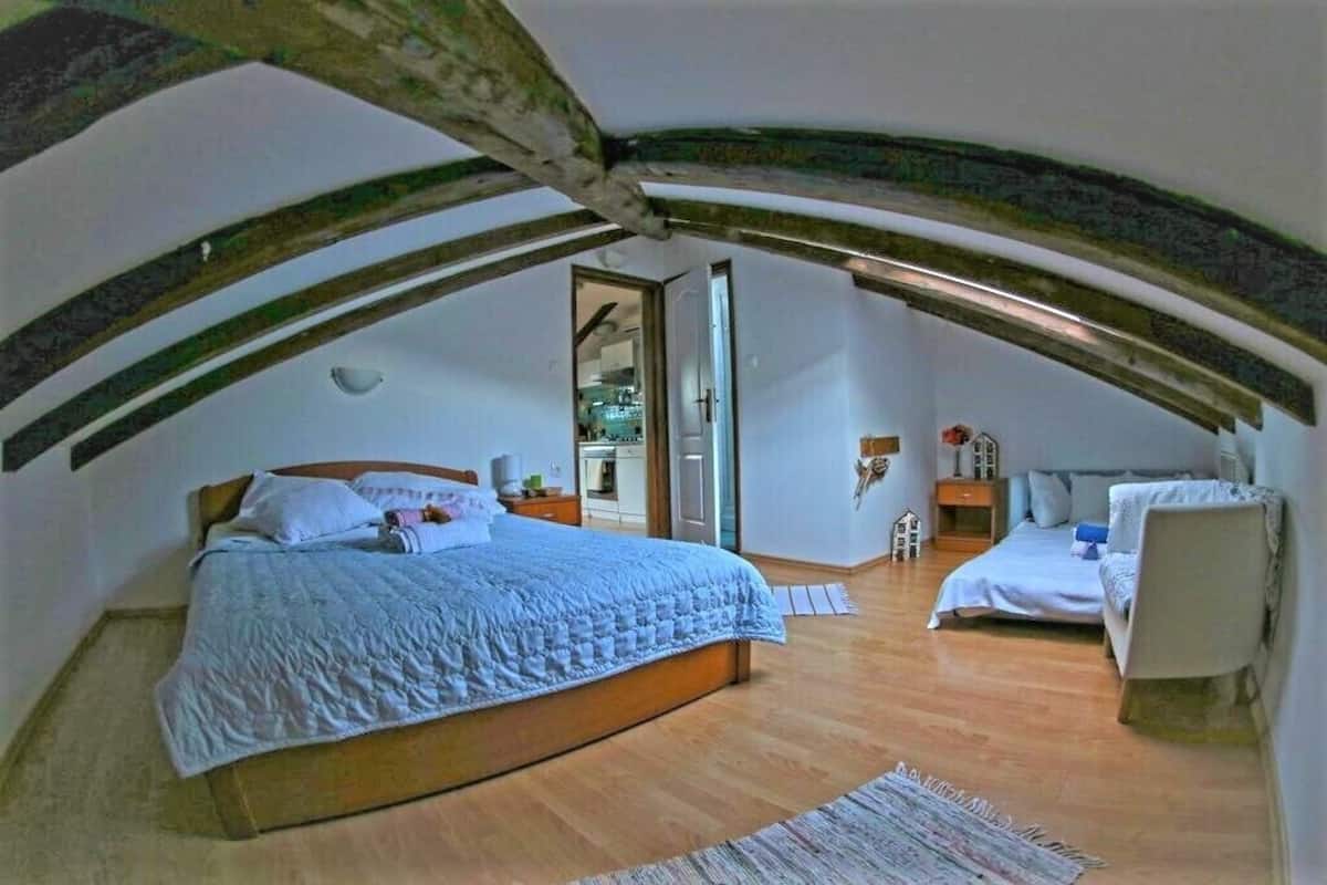 A bedroom with a large bed, a smaller bed, and a chair. There is a wooden ceiling with exposed beams.