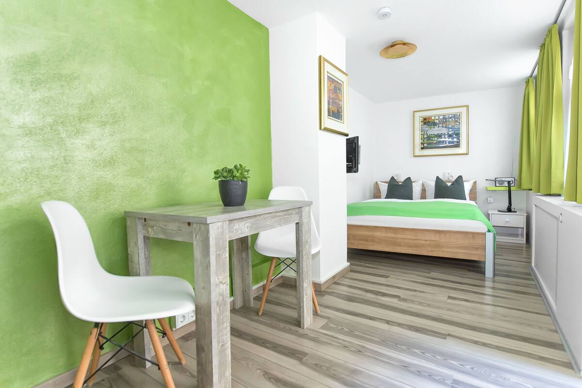 Ein modernes Zimmer mit grüner Akzentwand, einem hölzernen Bett mit grüner Bettdecke, einem weißen Stuhl, einem kleinen Tisch mit Topfpflanze und zwei gerahmten Bildern an der Wand.