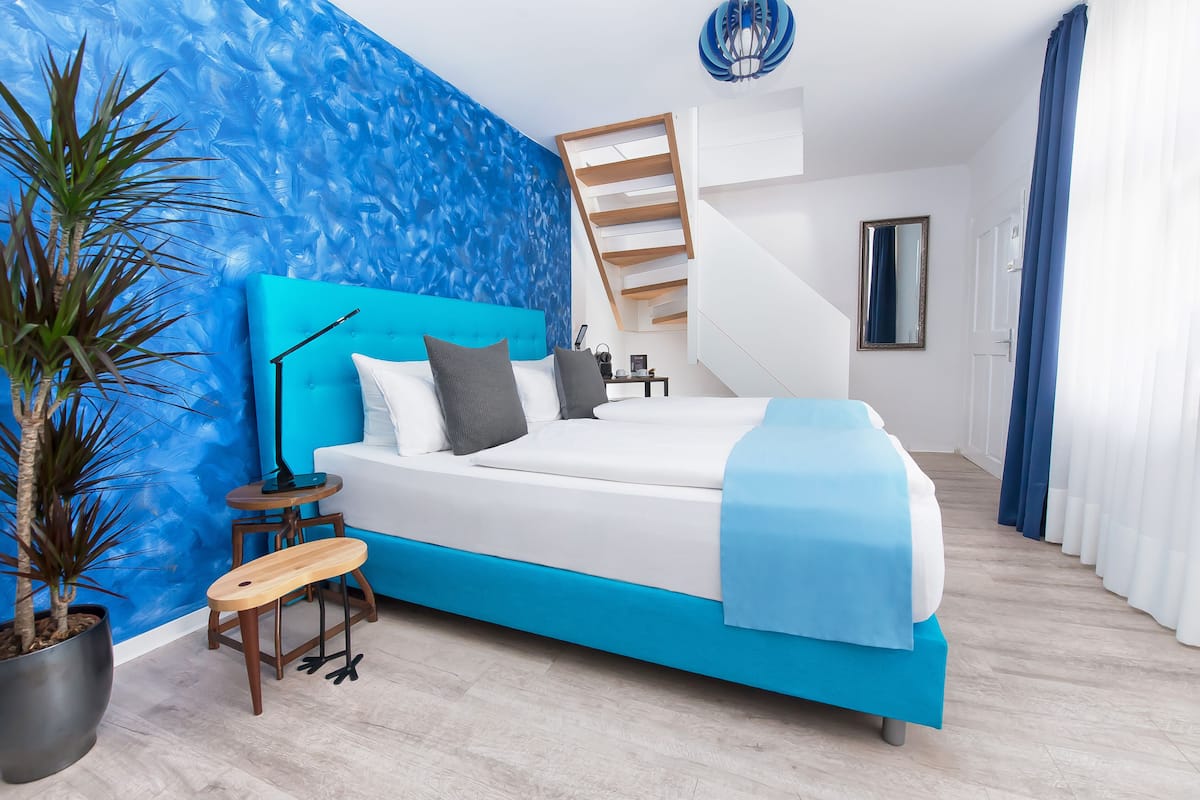 Ein modernes Schlafzimmer mit blauer Akzentwand, einem Bett mit blauer Kopfteil, einem kleinen Holzschemel, einem Topf mit Pflanze und einer Treppe.