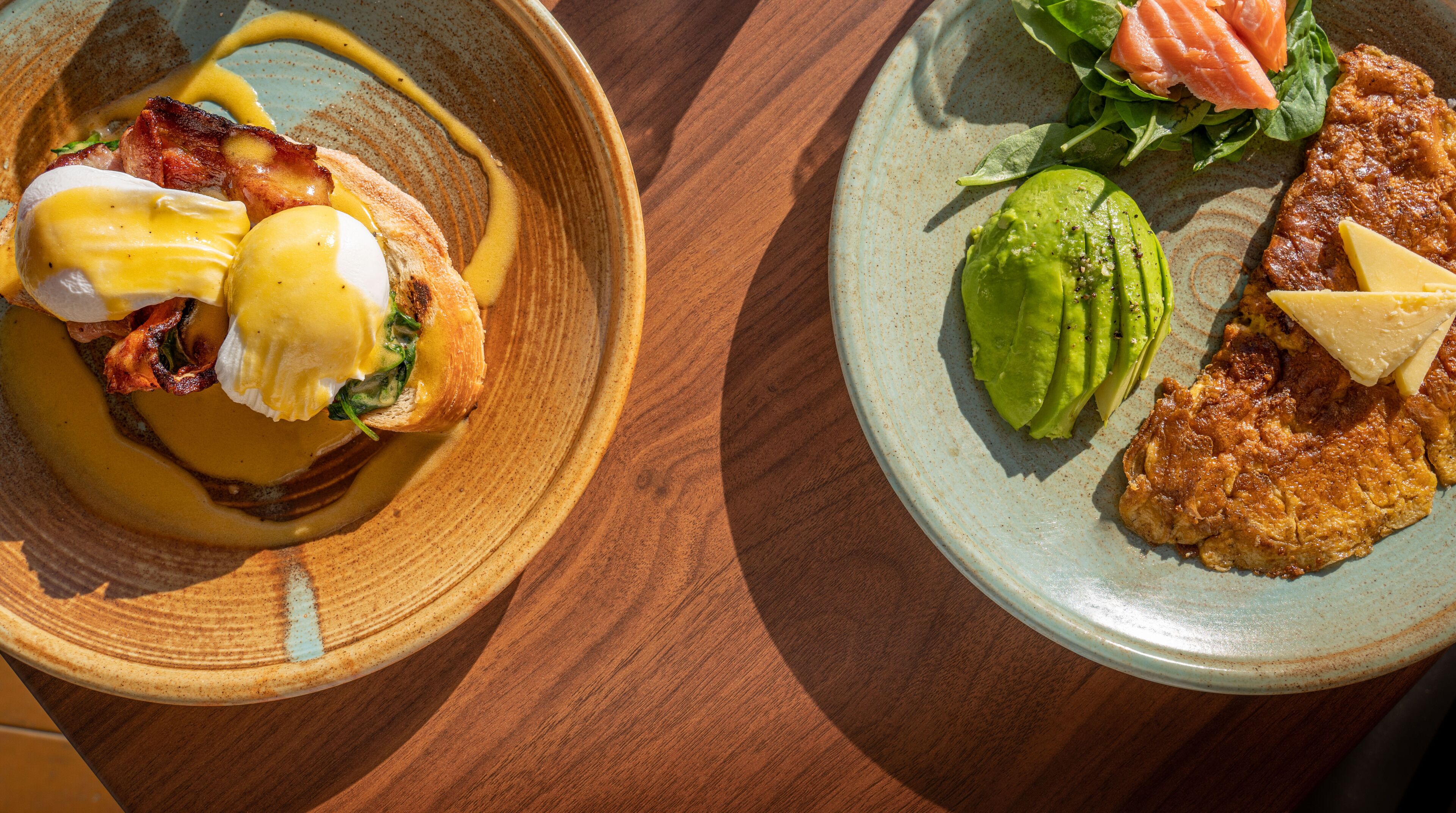 A wooden table with two plates of food, one with eggs Benedict and the other with a salad and avocado.