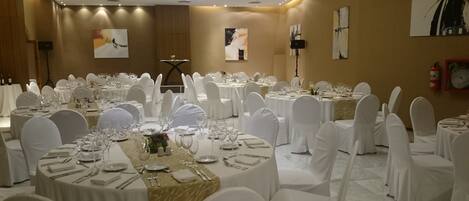 Un salón de banquetes con mesas redondas dispuestas para comer, sillas blancas y obras de arte en las paredes.