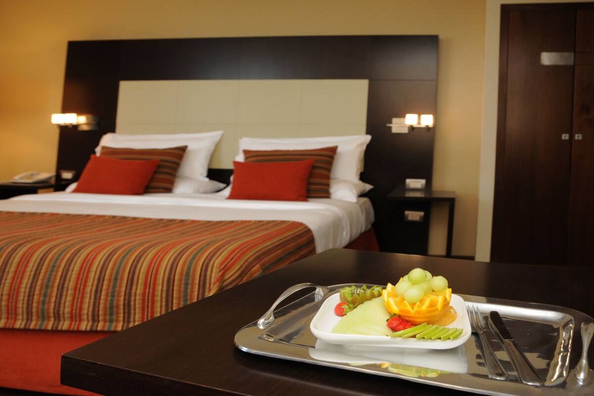 Une chambre d’hôtel avec un lit bien fait, un plateau avec des fruits et des couverts, et une table de chevet avec une lampe.