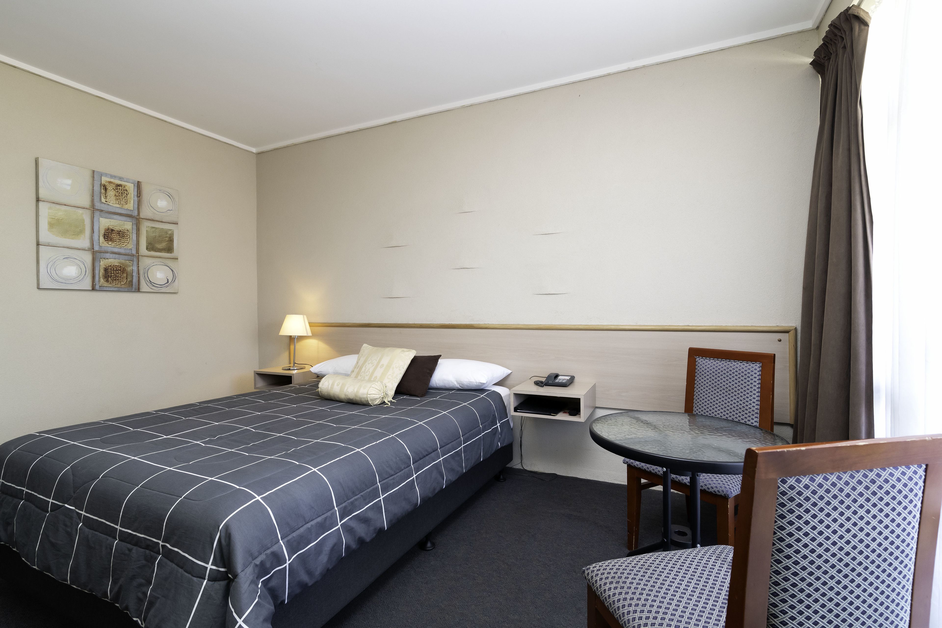 A hotel room with a bed, bedside table, lamp, chair, and a wall with artwork.