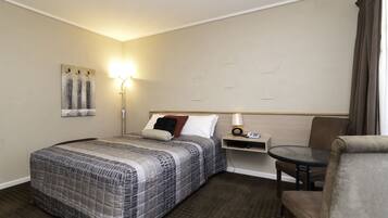 A hotel room with a bed, a chair, a small table, a lamp, and a wall-mounted artwork.