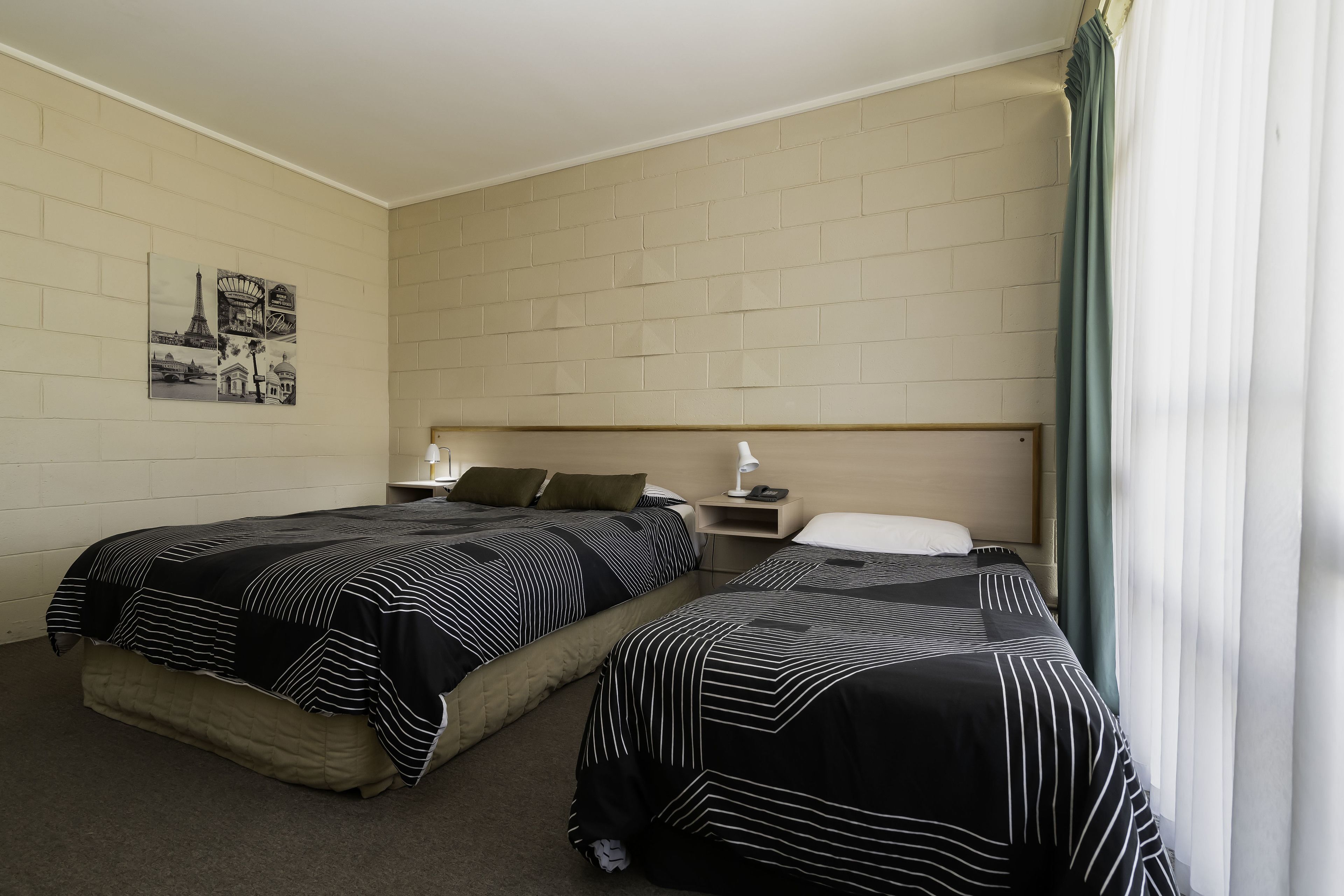 Une chambre d’hôtel avec deux lits, une table de chevet, une lampe et un mur orné d’un tableau représentant un paysage urbain.