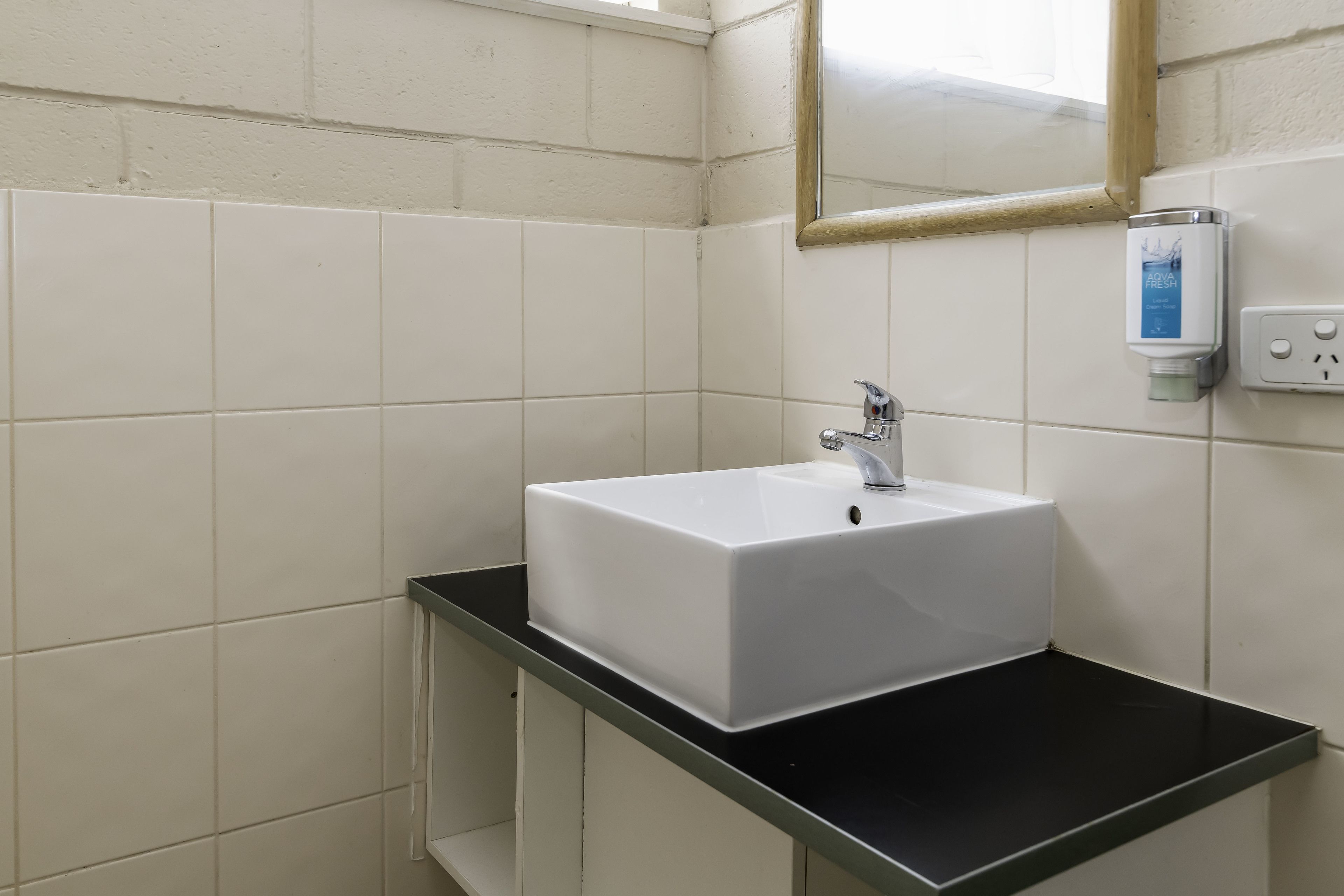 A bathroom with white tiles, a white sink, a black countertop, a mirror, and a hand sanitizer dispenser.