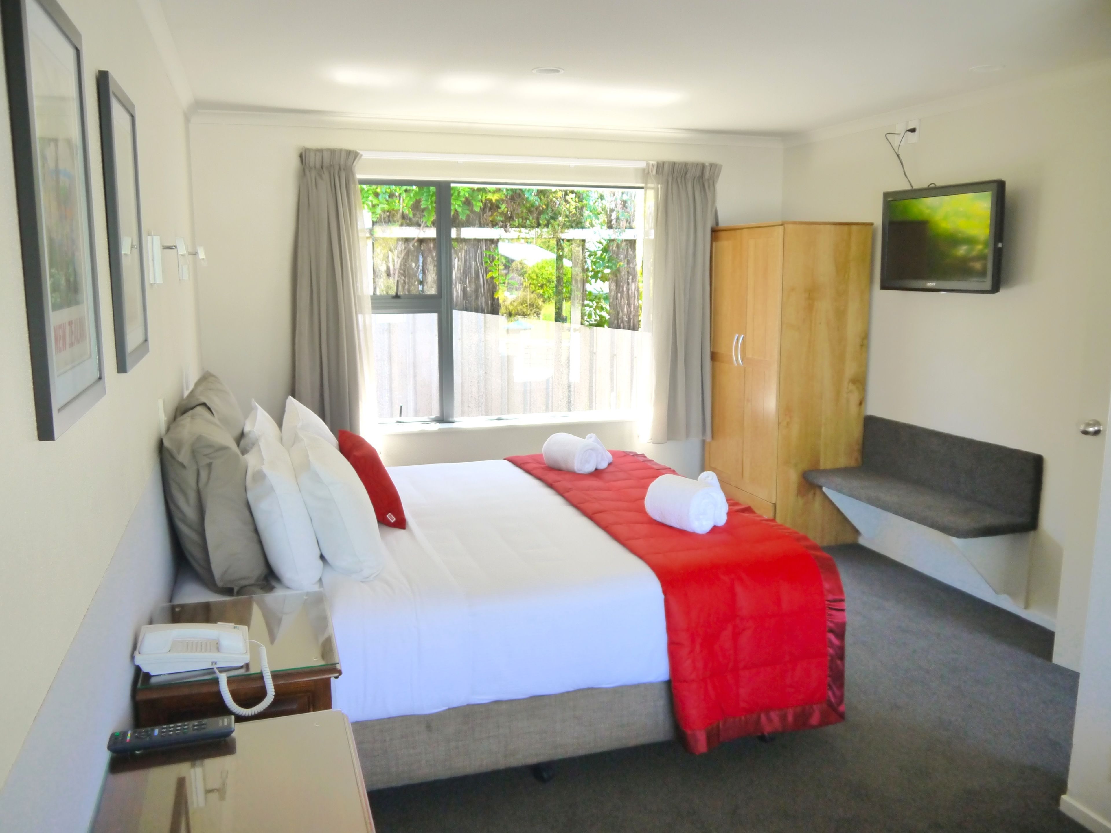 A hotel room with a bed, a desk with a telephone and remote control, a television on the wall, and a view of greenery outside.