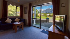 A living room with a sofa, a coffee table, a TV, and a large sliding glass door leading to a balcony with a mountain view.