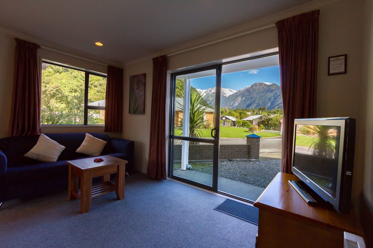 A living room with a sofa, a coffee table, a TV, and a large sliding glass door leading to a balcony with a mountain view.