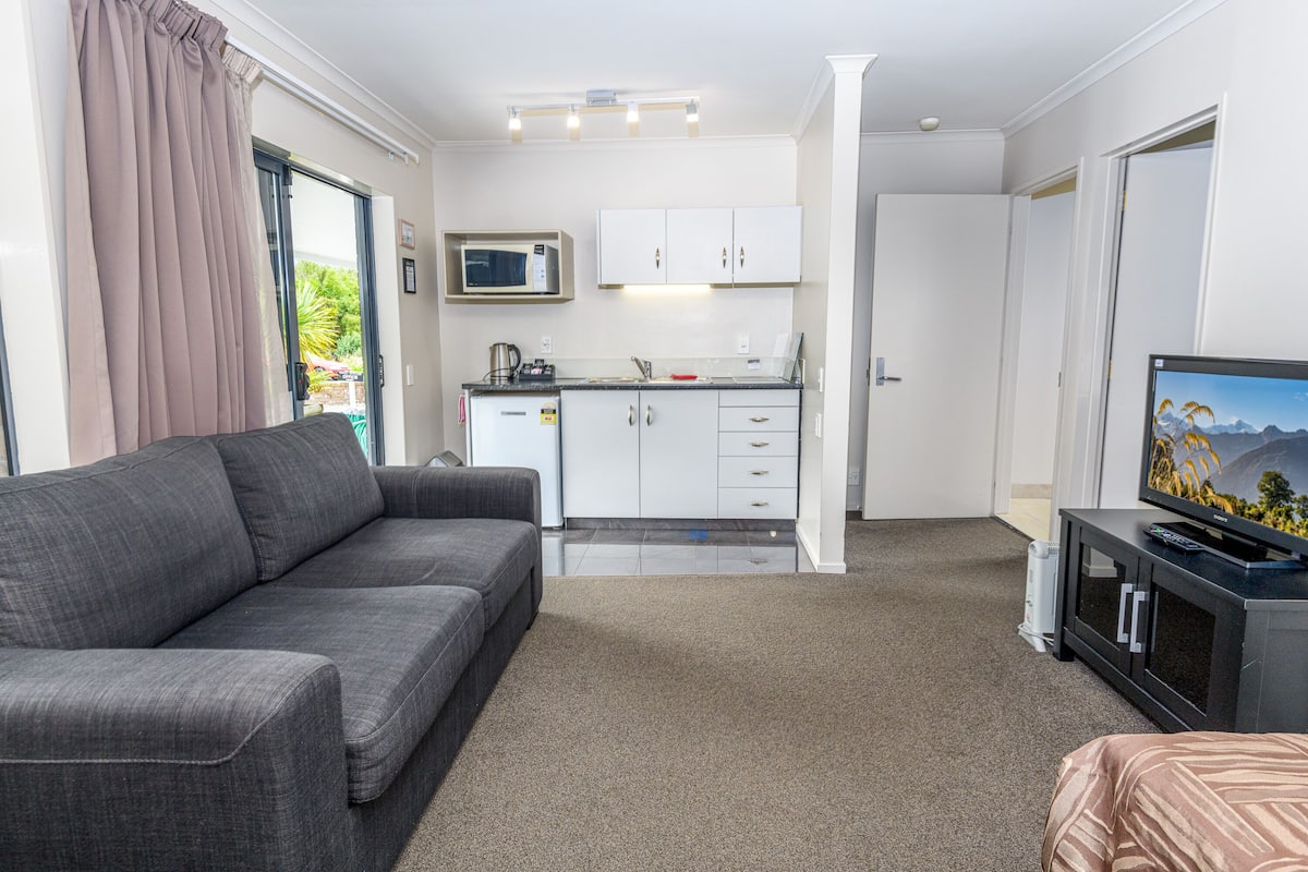 A modern hotel room with a grey sofa, a television, a kitchen area with white cabinets, and a carpeted floor.