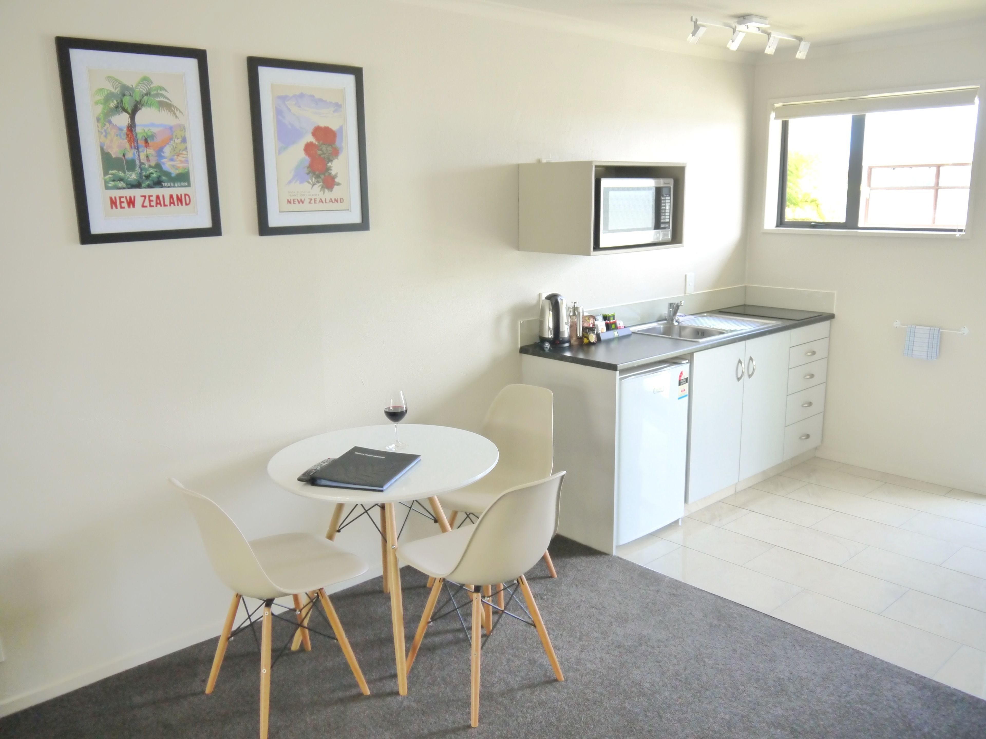 Une cuisine moderne équipée d’un four à micro-ondes, d’un évier et de placards. Un coin repas avec une table et des chaises est également disponible. Deux tableaux encadrés représentant des paysages de Nouvelle-Zélande ornent le mur.