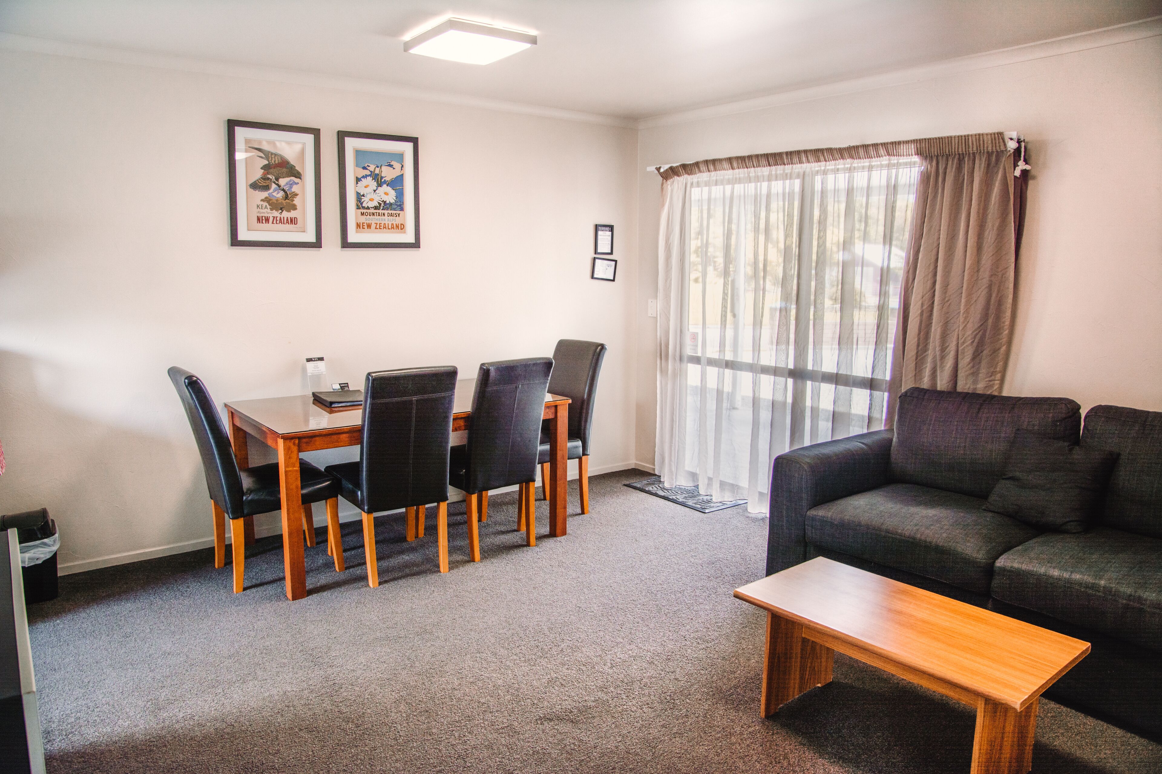 A living room with a sofa, a coffee table, and a dining area with a table and chairs. There are two framed pictures on the wall.