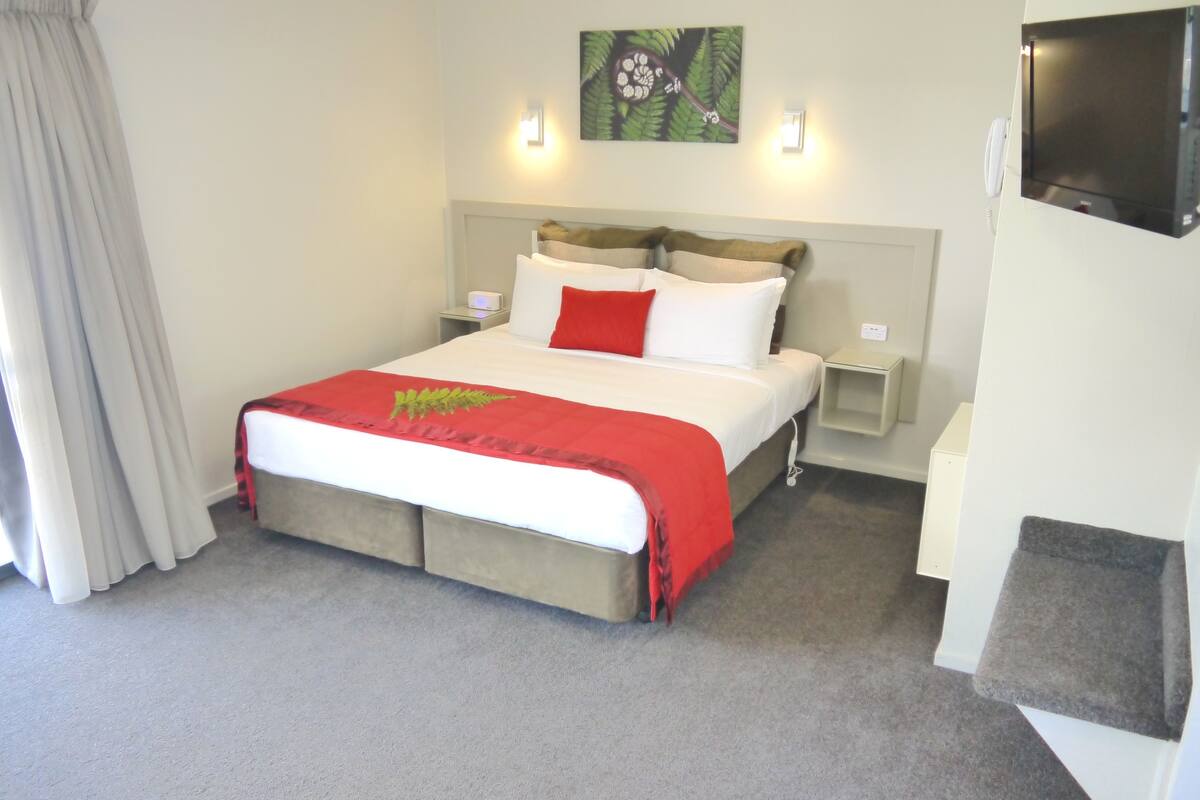 A hotel room with a bed, a nightstand, a TV mounted on the wall, and a painting above the bed.