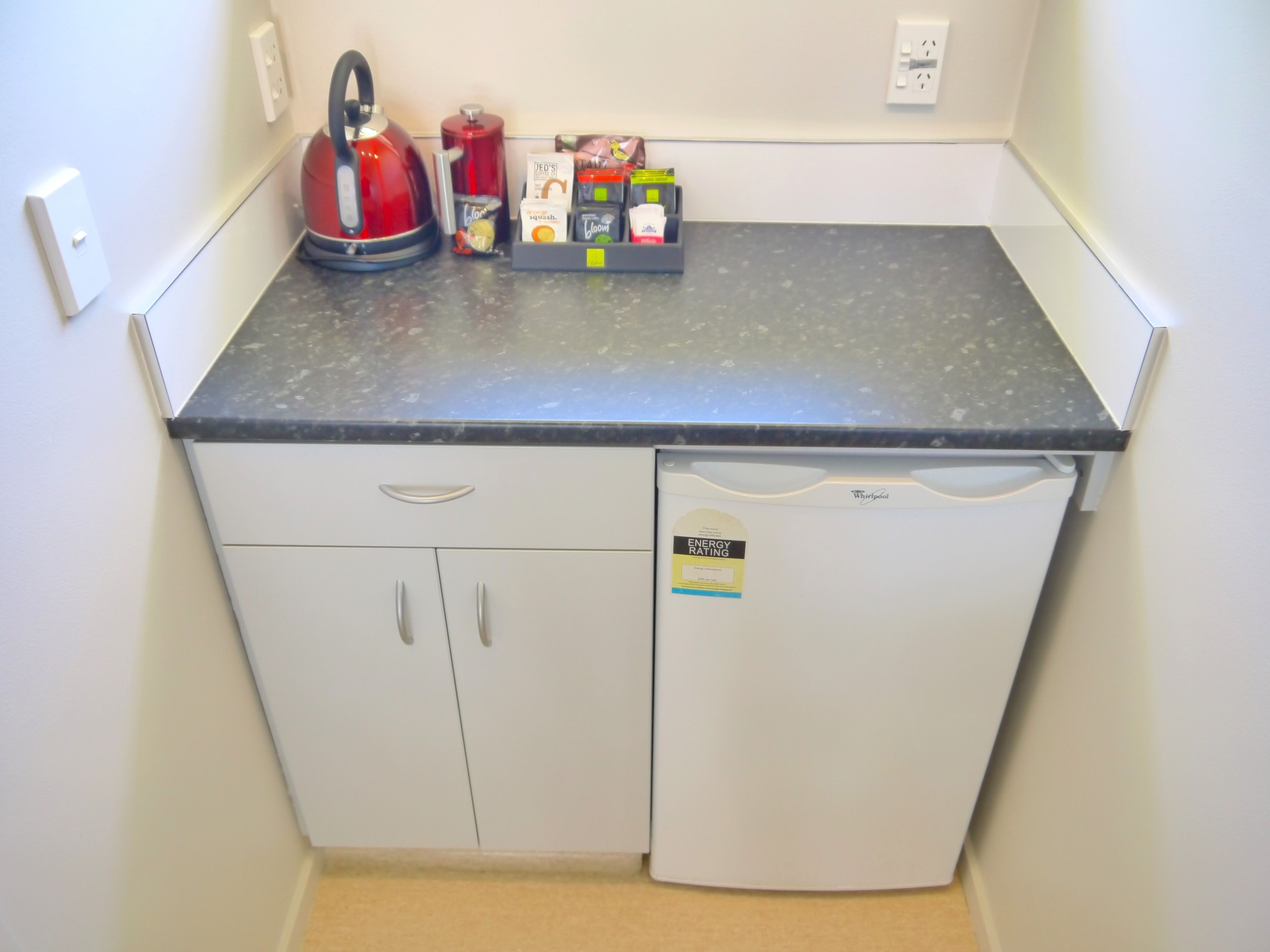 A kitchenette with a black countertop, a red kettle, a coffee maker, and a small refrigerator.