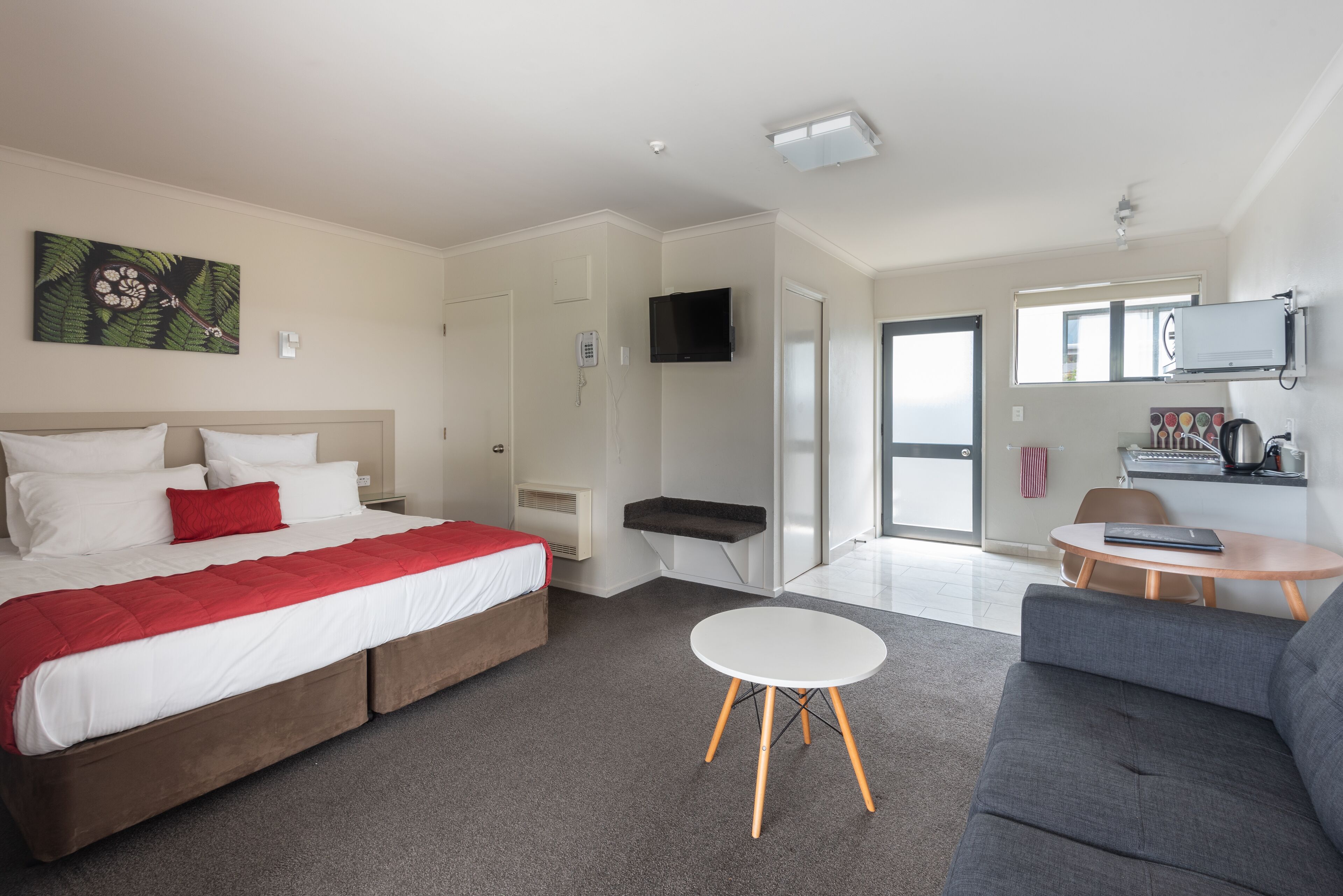 A modern hotel room with a large bed, a grey sofa, a small round table, and a TV mounted on the wall.