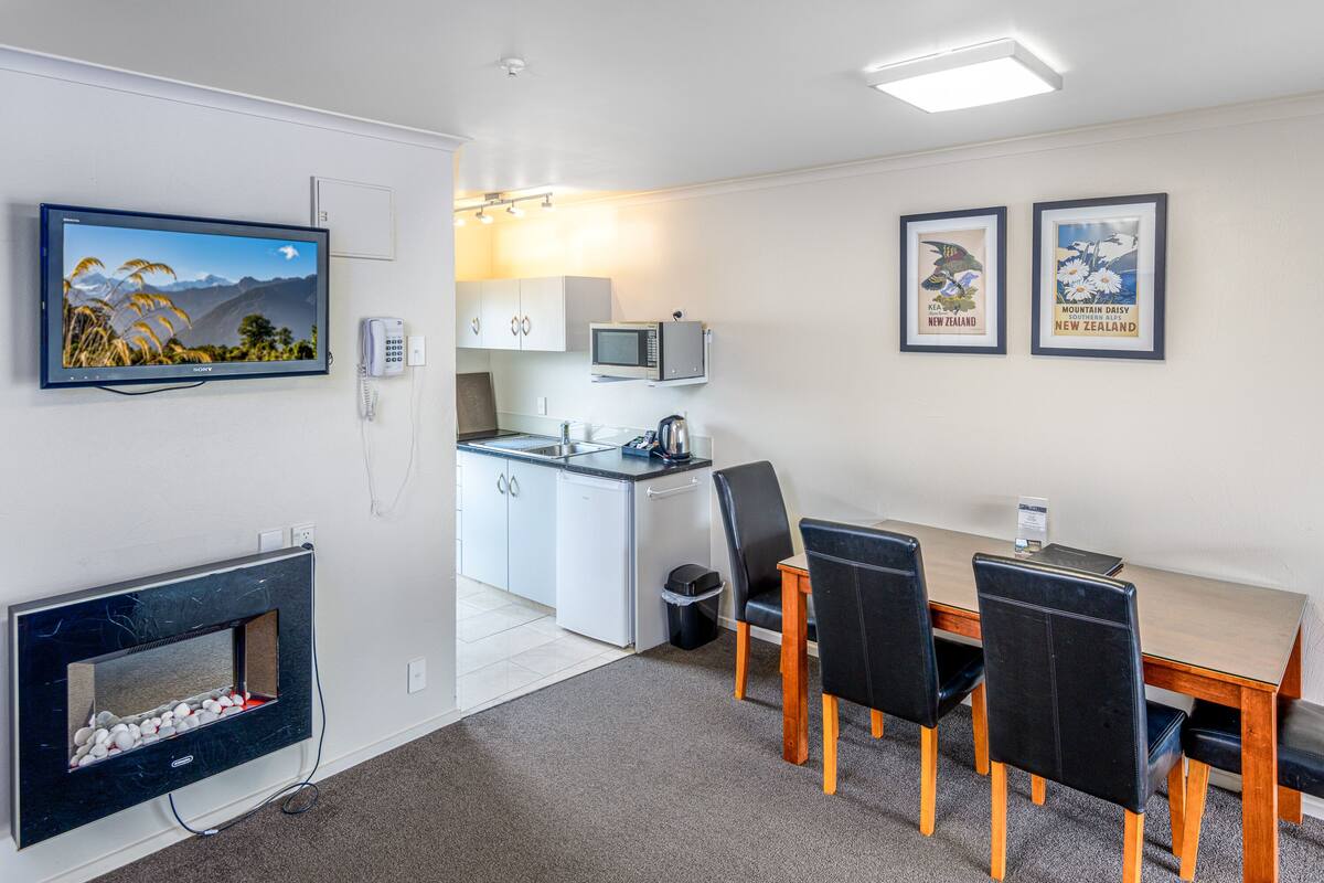 A compact kitchen with a microwave, a small refrigerator, and a sink. There is a dining table with four chairs and a flat-screen TV mounted on the wall. Two framed pictures are displayed on the wall.