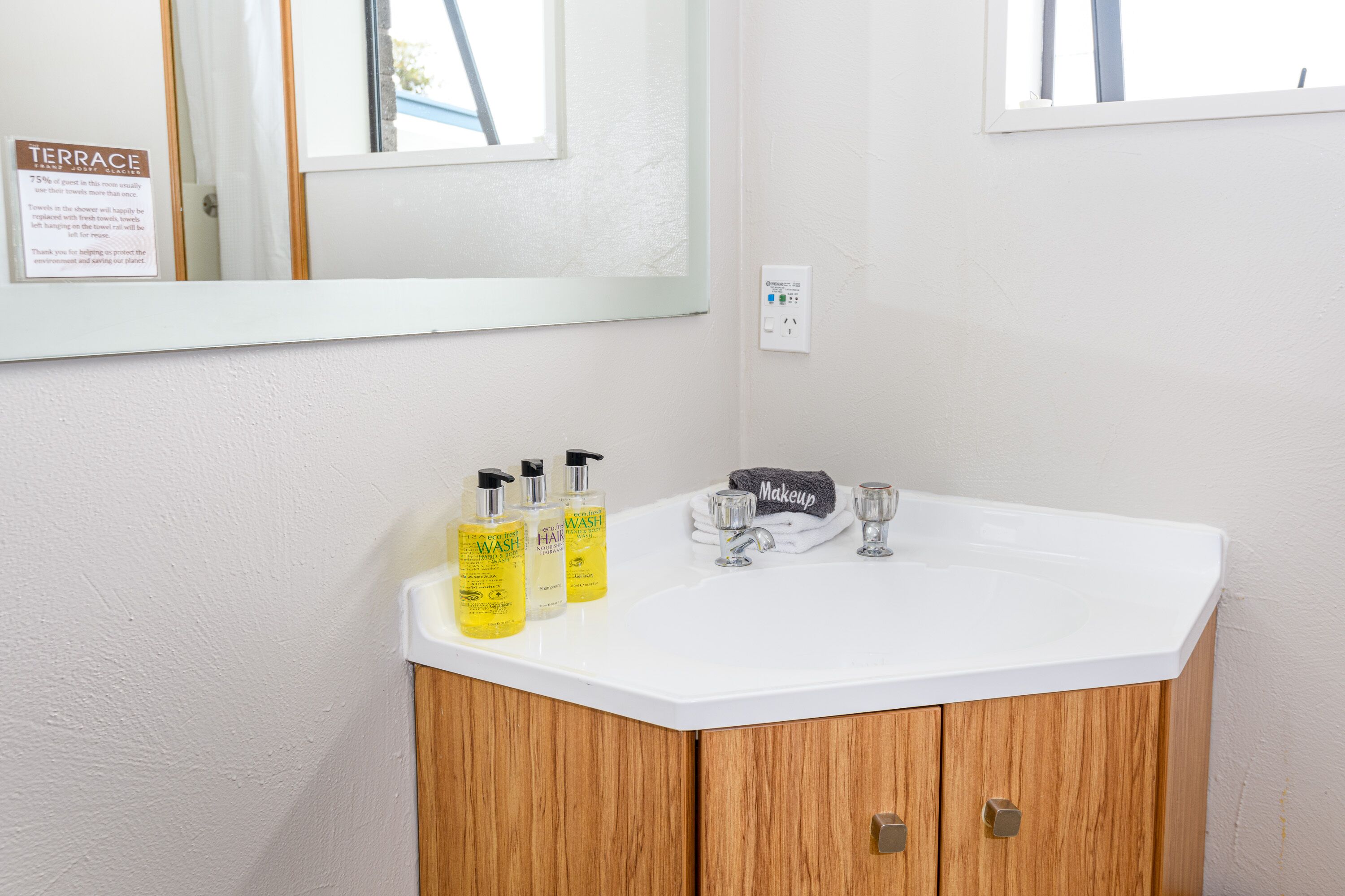 Une salle de bain avec un lavabo blanc, des meubles en bois et un miroir. On y trouve trois bouteilles de shampoing et d’après-shampoing posées sur le lavabo.