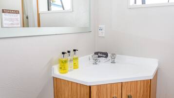 Une salle de bain avec un lavabo blanc, des meubles en bois et un miroir. On y trouve trois bouteilles de shampoing et d’après-shampoing posées sur le lavabo.