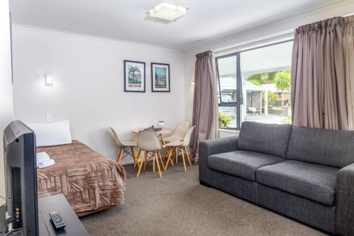 A hotel room with a bed, a sofa, a TV, a desk with chairs, and a window with curtains.