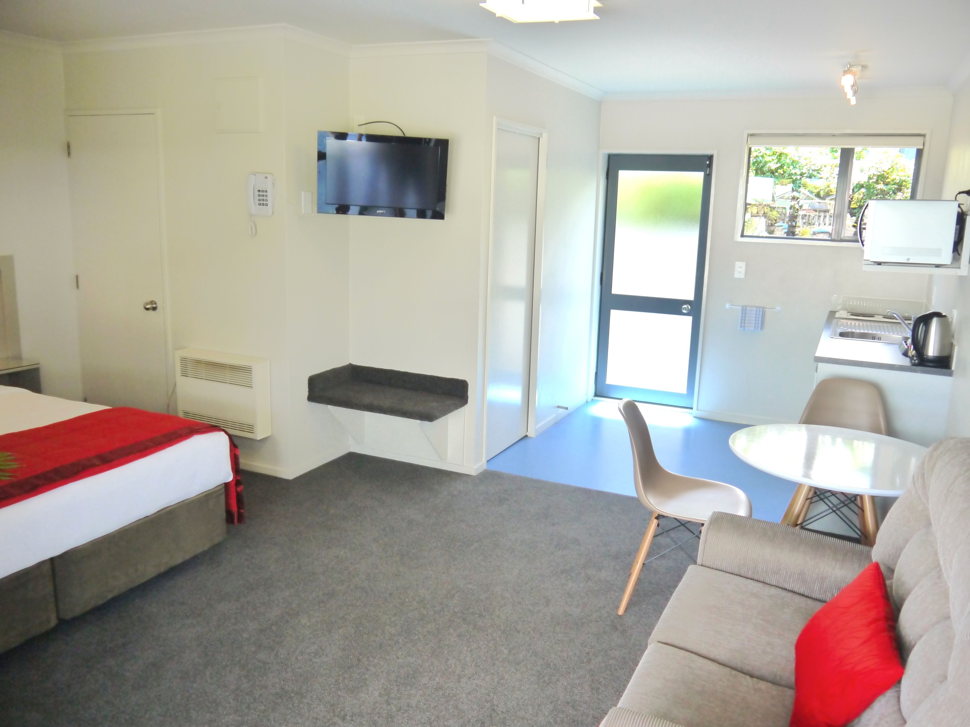 A hotel room with a bed, a sofa, a small table, a TV mounted on the wall, and a door leading outside.