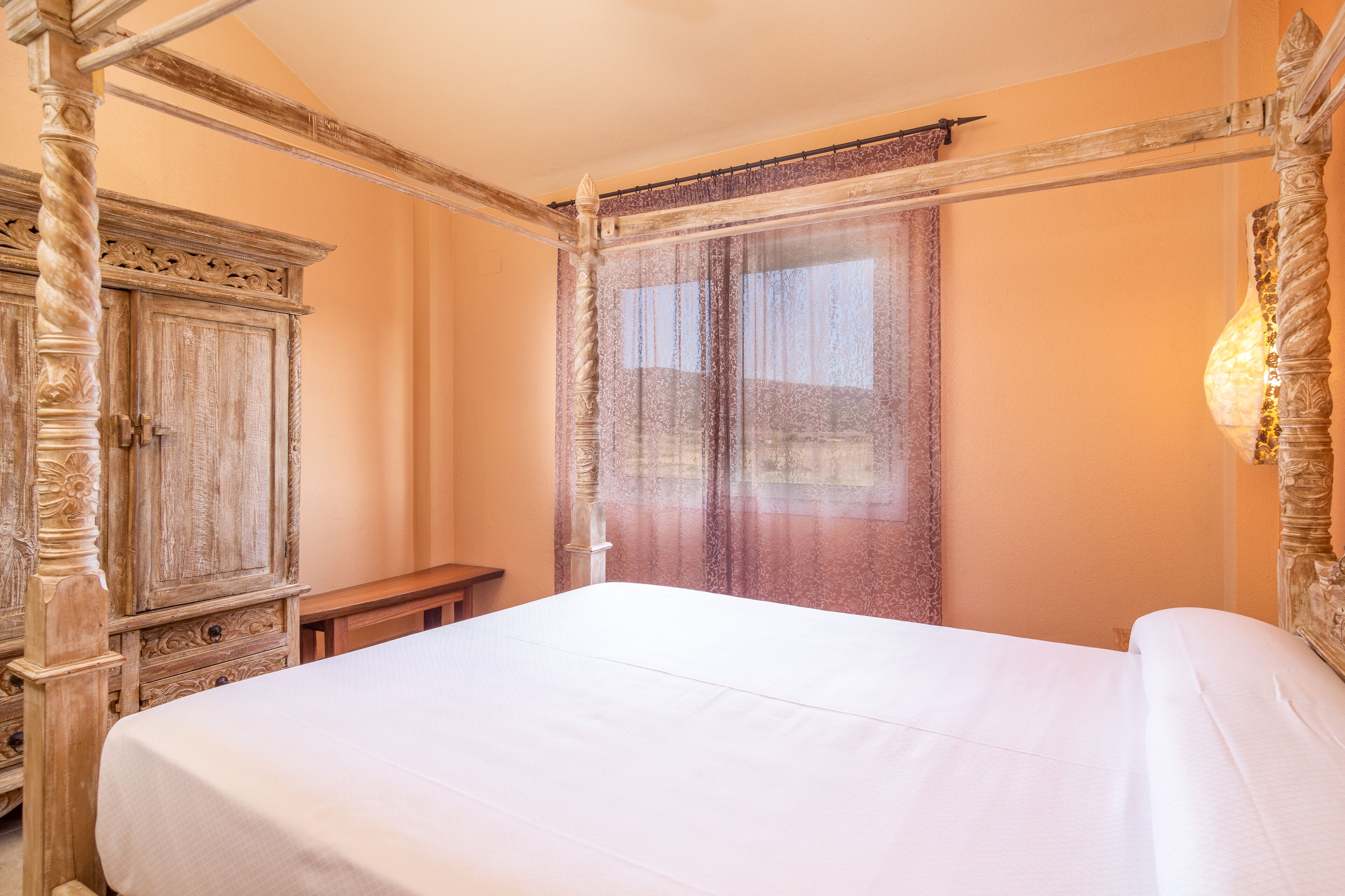 Un lit à baldaquin avec une couvre-lit blanche, une armoire en bois et une fenêtre ornée de rideaux légers.