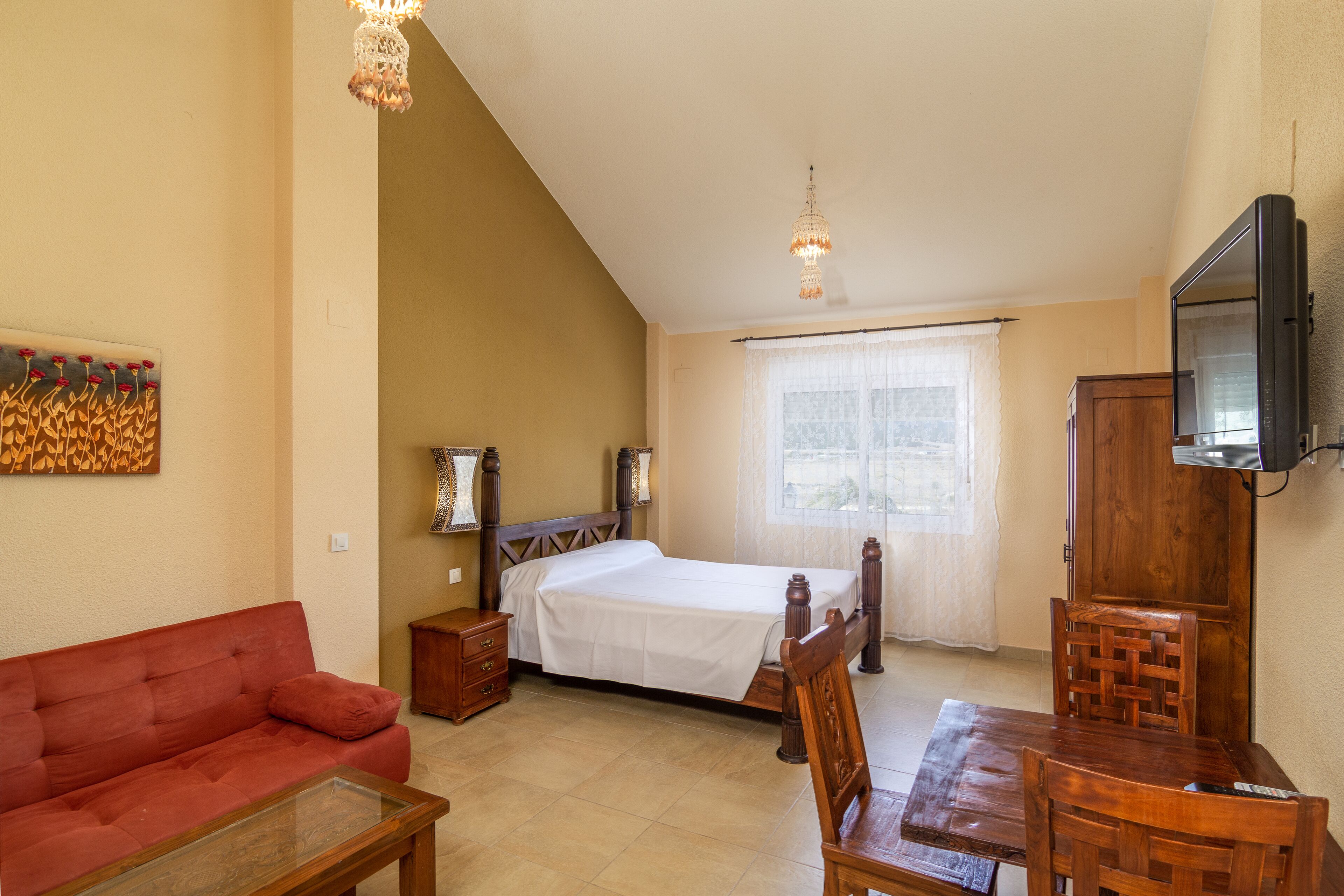 A bedroom with a bed, a wooden table, a red sofa, a TV mounted on the wall, and a window with curtains.