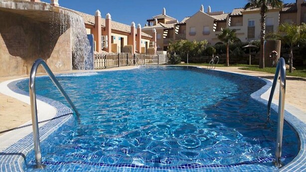 Una piscina con agua azul cristalina, rodeada por una solana con baldosas y un muro de piedra.