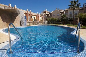 Una piscina con agua azul cristalina, rodeada por una solana con baldosas y un muro de piedra.