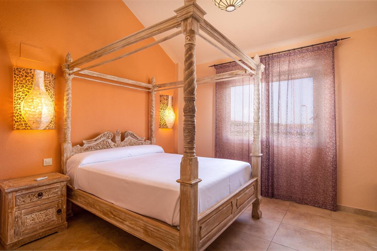 A four-poster bed with a wooden frame and white bedding, a nightstand with a drawer, and a window with a purple curtain.
