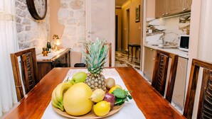 A dining table with a fruit platter, wooden chairs, a stone wall, a clock, and a kitchen in the background. - D & Sons Apartments (Kotor)