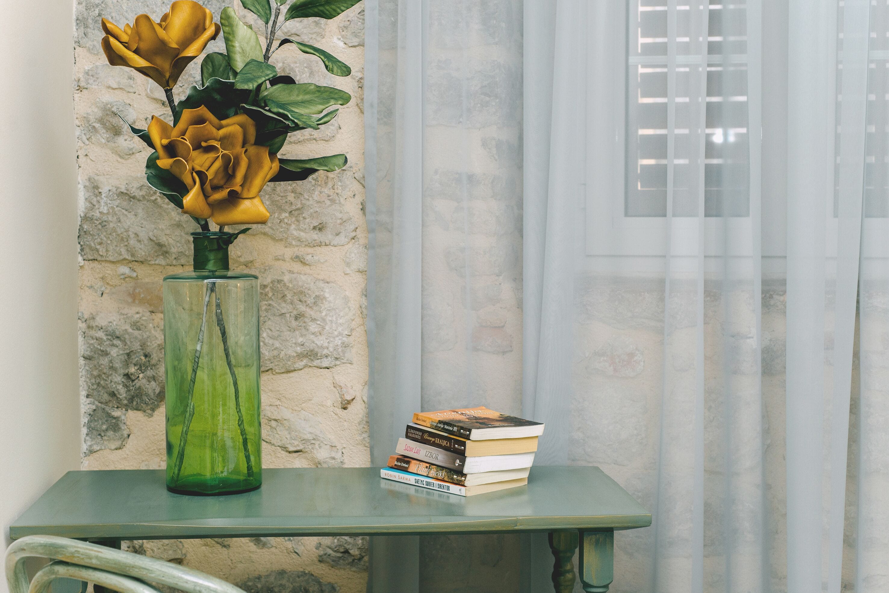 A green vase with yellow flowers on a glass table next to a stack of books.
